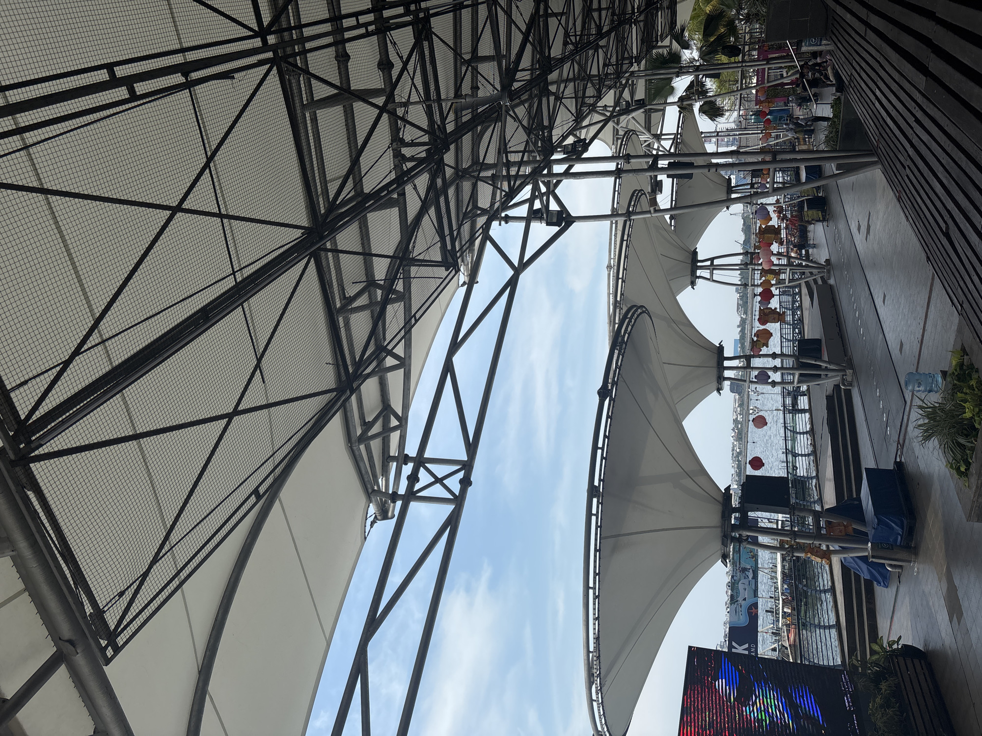 The image depicts an outdoor space in North Jakarta, Indonesia, covered by a large, white canopy supported by metal beams and scaffolding. The sky is visible between the canopies, showing light blue with scattered clouds. Hanging decorations, possibly lanterns, are strung beneath the canopies. Below, the space features a walkway and steps leading down to a waterfront area where boats are visible. A large electronic screen displays colorful imagery. The overall impression is that of a public area designed for events or gatherings.