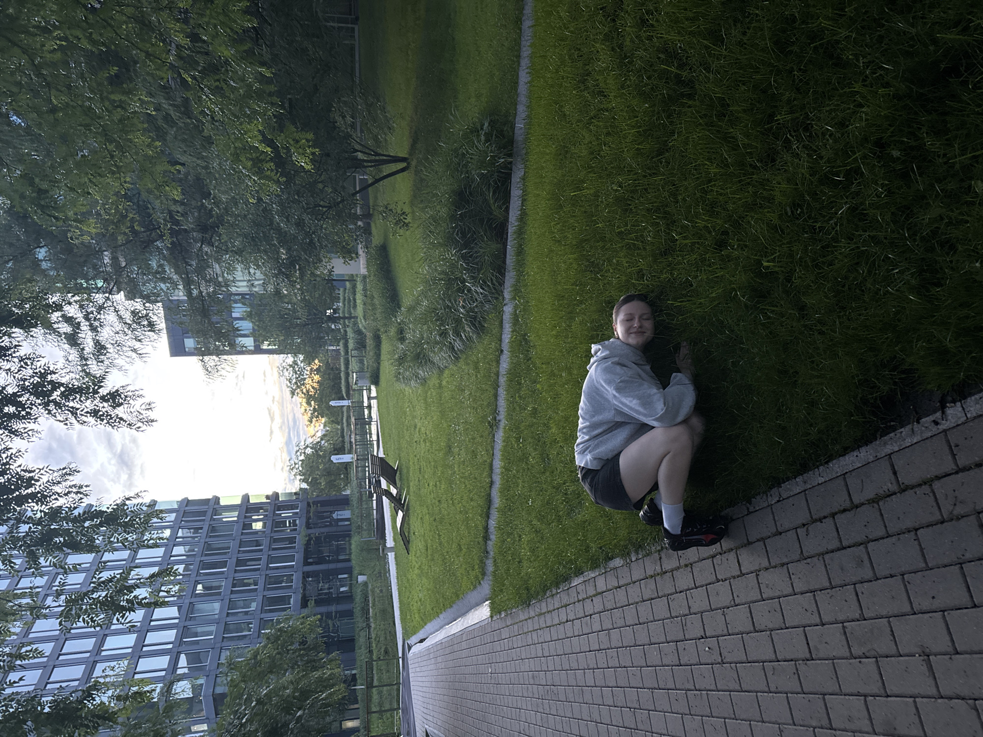 An overhead shot shows a person lying on their side in lush green grass next to a brick pathway. The person is wearing a grey hooded sweatshirt, black shorts, white socks, and black sneakers with red accents. Their head is resting on their arm, and they are smiling. In the background, two modern glass buildings stand tall, surrounded by trees and a cloudy sky. A park-like setting with benches and manicured greenery is visible between the buildings. The lighting suggests it is either early morning or late evening.
