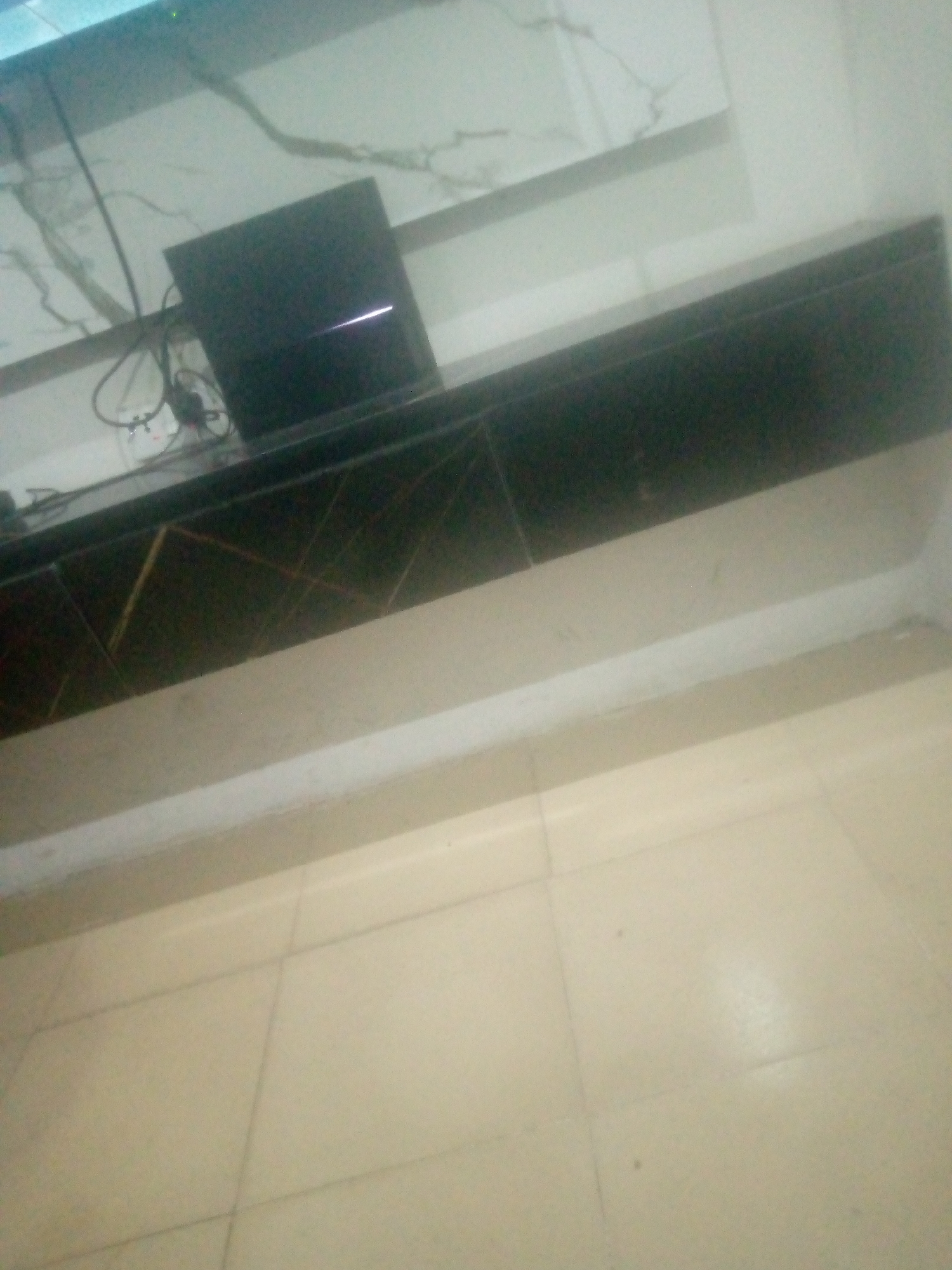 A black electronic device, possibly a game console, sits on a dark, reflective surface. The surface appears to be a shelf or cabinet, with a marbled white and grey design on the wall behind it. The floor is tiled with light brown squares. The scene is indoors, with no indication of the specific location within Jalingo, Nigeria, beyond it being an interior space. The lighting is dim, and the image quality is poor, making it difficult to discern further details about the device or its surroundings.