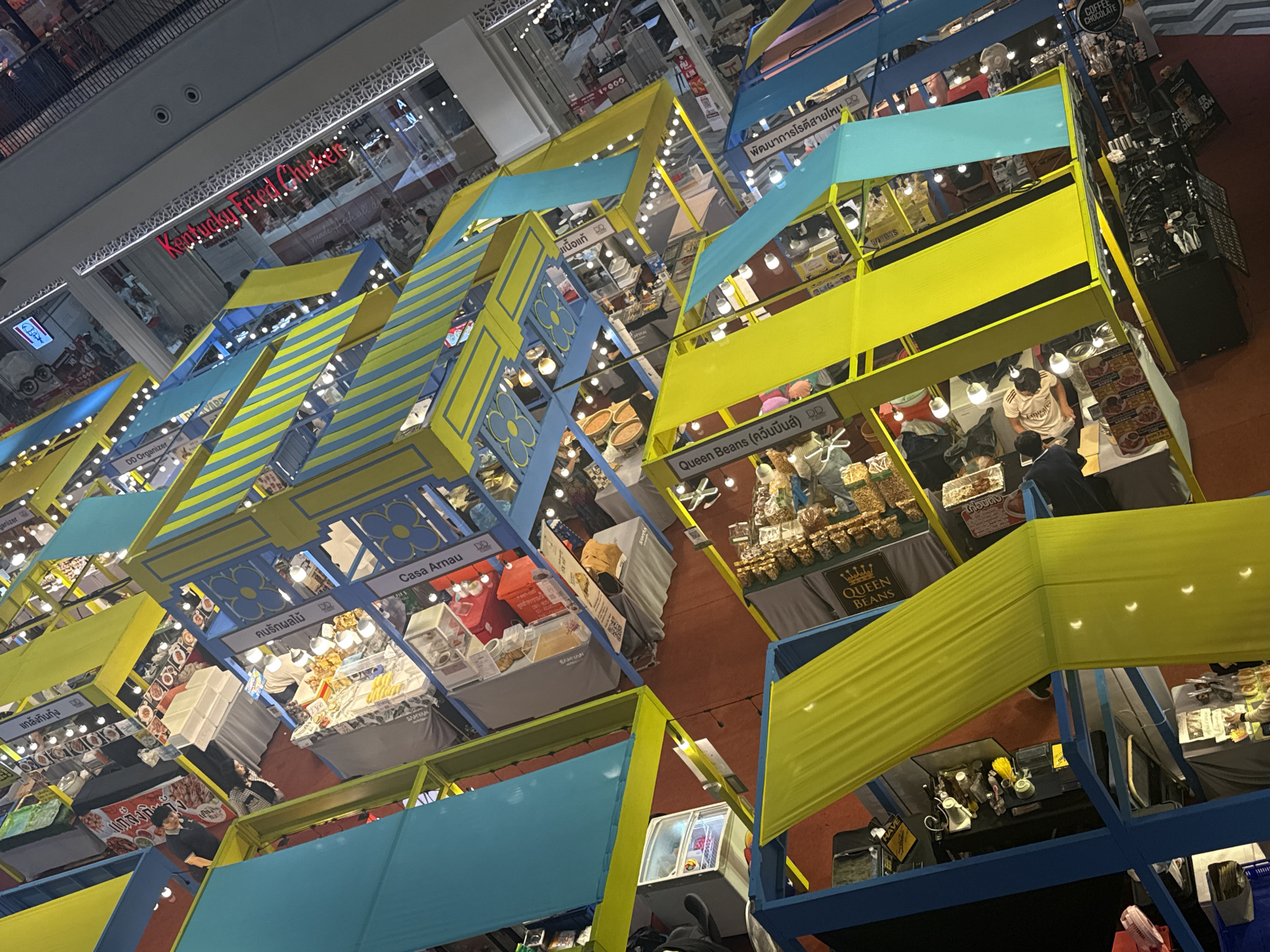 The image depicts a bustling indoor market or exhibition hall, likely in Bangkok, Thailand, given the signage in Thai script visible on some of the stalls. The scene is characterized by numerous vendor stalls with colorful awnings, primarily in bright yellow, blue, and teal. These stalls are set up in a dense arrangement, forming aisles and creating a vibrant, somewhat chaotic atmosphere.
Several stalls display various goods, including food items, baked goods, and possibly packaged products. Some stalls have product displays on tables, while others feature refrigerators or glass display cases. People are seen browsing and interacting at the stalls, with some vendors attending to customers. The lighting appears to be a mix of overhead artificial lights and individual stall lighting.
The overall perspective is from an elevated angle, looking down into the market space. The architecture of the building is visible in the background, with elements like railings, upper floors, and structural supports suggesting a large commercial or exhibition venue. The floor of the market appears to be a reddish-brown material. The image captures a lively scene of commerce and public activity within an indoor marketplace.