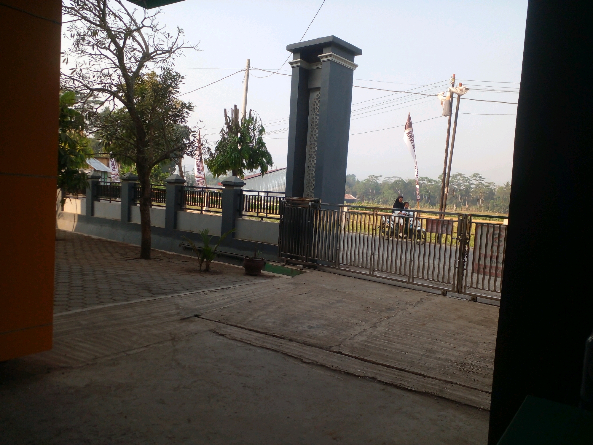 In Banyuputih, Indonesia, a street scene unfolds with a paved walkway in the foreground leading to a metal gate. To the left, an orange building with a dark tiled wall provides a backdrop. A young tree stands near the walkway, and a potted plant sits beside it. Beyond the walkway, a dark gray rectangular pillar with decorative cutouts marks the entrance. A metal fence runs along the property line, separating it from a road where two individuals on a motorcycle are visible. The motorcycle is turning into the property. In the background, a field of rice paddies stretches towards a treeline under a clear sky. Flags fly on poles, and utility lines crisscross the sky.