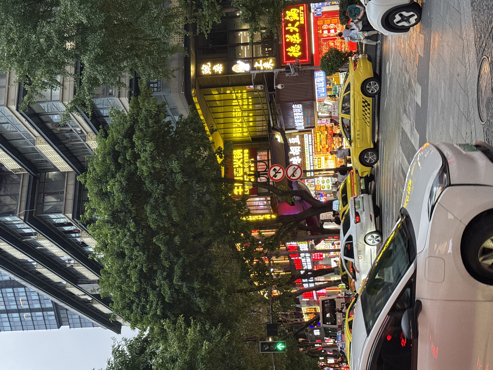 The image depicts a street scene in Yuzhong District, China, on what appears to be a cloudy day, given the ambient lighting. The foreground is dominated by the front and side of a white car, suggesting it is either stopped or moving slowly. Behind it, a yellow taxi is visible, with its driver's arm resting on the open window. Several other vehicles, including a white sedan and a bus, are present on the street, indicating moderate traffic.
The background is characterized by a multi-story building adorned with numerous neon signs in Chinese characters, many of which are illuminated, casting a glow on the street. These signs suggest a commercial area, possibly with restaurants and shops. Trees line the street, their dense foliage partially obscuring the buildings and signs. A traffic light with a green arrow is visible on the left side of the image. A few pedestrians are scattered along the sidewalk and street, engaged in various activities like sitting or walking. The overall impression is a busy urban environment with a vibrant streetscape at dusk or on an overcast day.
