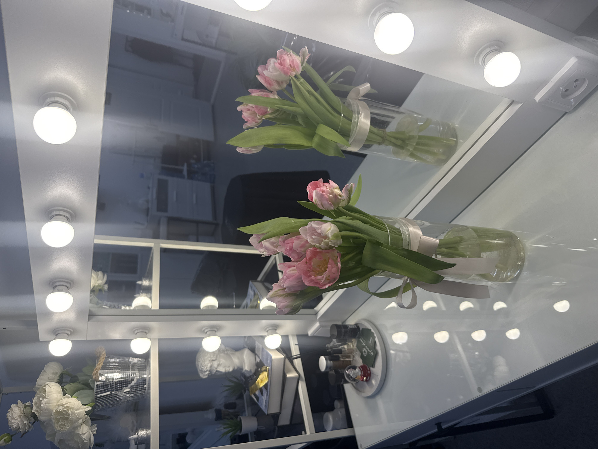 The image depicts a mirrored vanity setup, likely in a room within a dwelling in Chaikovsky, Russia. The vanity is illuminated by multiple white light bulbs arranged around the perimeter of the mirror. On the table, two glass vases containing pink and white tulips are visible. The mirror reflects the room behind the viewer, showing details of the interior, including a partially visible disco ball, a bust of a man, plants, and miscellaneous items on the vanity’s surface. There are no people in the image.