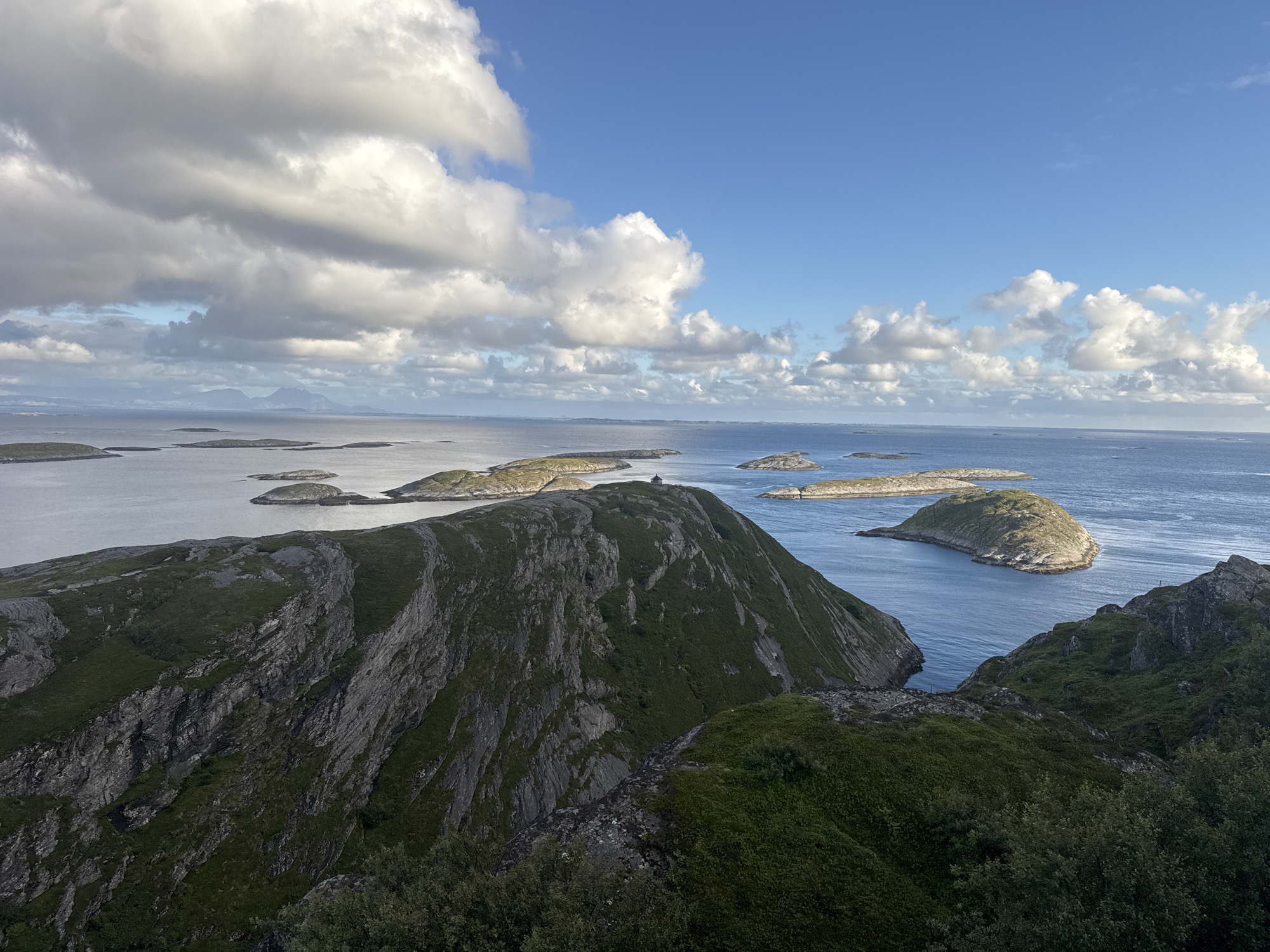This is an eye-level, wide shot photograph taken from a high vantage point, looking out over a coastal landscape. The foreground is dominated by a steep, rocky, and vegetated hillside. The middle ground features several small, rocky islands scattered across a calm, blue body of water. In the distance, more islands and a hazy mountainous coastline are visible. The sky is a vibrant blue, with scattered, fluffy white clouds and larger, darker clouds casting shadows.
The specific location is the summit of Lovund mountain on Lovund island in Norway. A small, dark-colored building with a steep roof is visible on the crest of one of the prominent islands in the middle distance. The building appears to be a small house or shelter. The rugged terrain of the foreground is characterized by dark gray rock faces interspersed with patches of green grass and low-lying vegetation. The islands are covered with similar vegetation and appear to be uninhabited except for the structure on one. The water is a deep blue, reflecting the sky and clouds. The overall impression is one of a serene, wild, and remote natural environment.