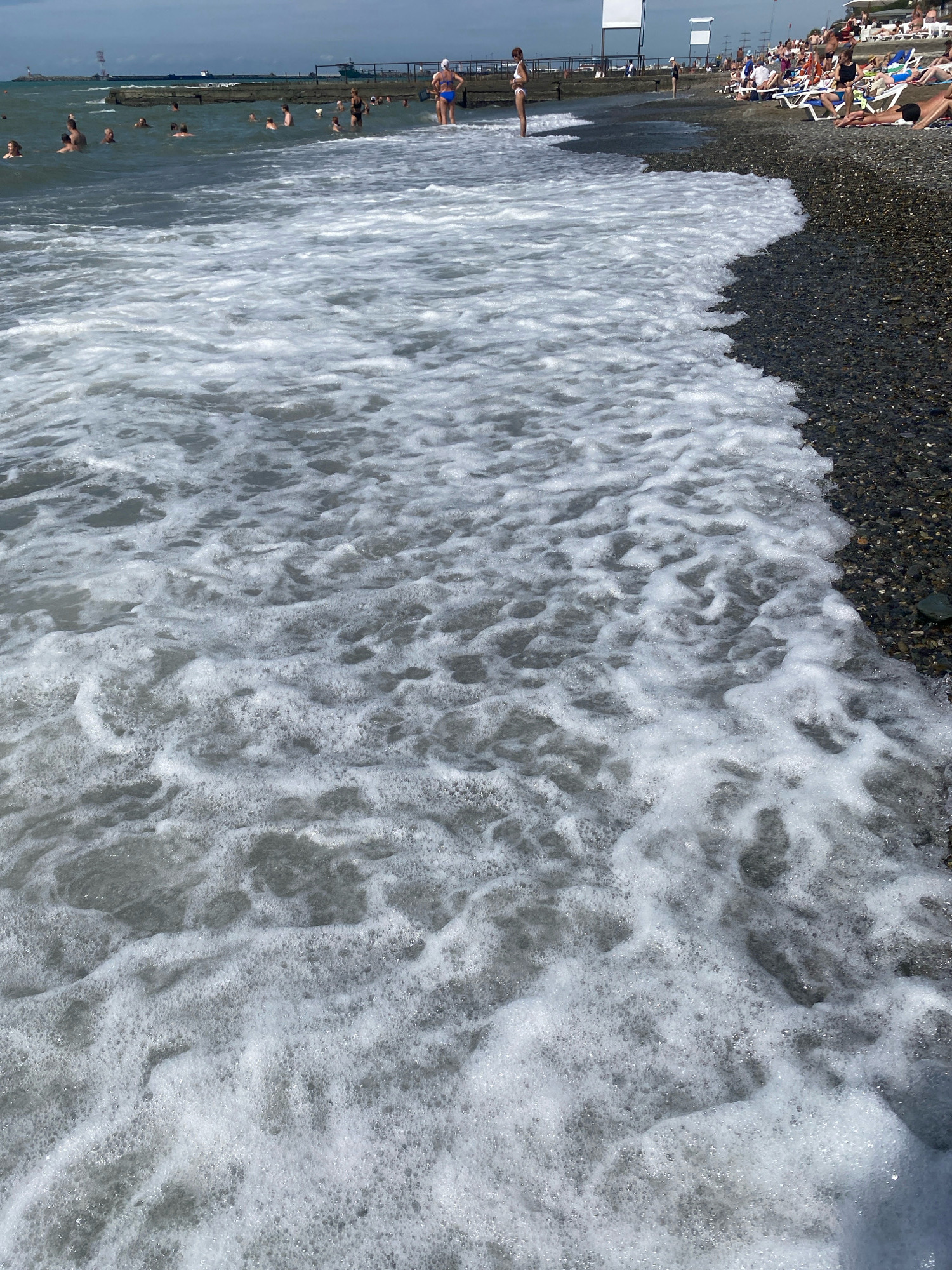 The image depicts a crowded beach scene on a sunny day, likely in Sochi, Russia, given the presence of sunbathers, umbrellas, and the characteristic pebble beach. The foreground is dominated by foamy seawater washing ashore, with numerous people visible in the background swimming, wading, and relaxing on lounge chairs. A pier or breakwater extends into the sea, with a ship visible in the distance. The sky is partly cloudy.