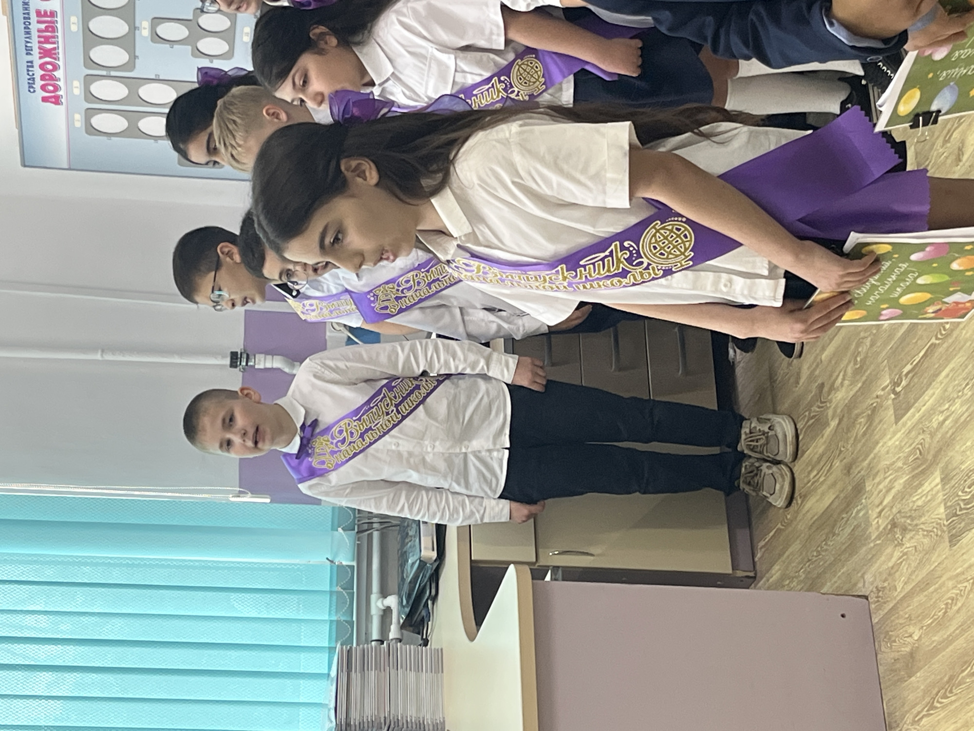 A photograph captures a group of students, presumably in Nizhnyaya Shilovka, Russia, possibly celebrating an occasion like graduation. The students are standing in a row, wearing white shirts and dark trousers/skirts, each adorned with a purple sash. The sashes feature a circular emblem and Cyrillic text, suggesting the end of primary school.
The background includes a wall with a poster displaying diagrams, a pale purple wall, and a window covered with light blue blinds. A desk is visible in the foreground.
The students appear to be holding books or certificates. The lighting suggests an indoor setting during daylight.