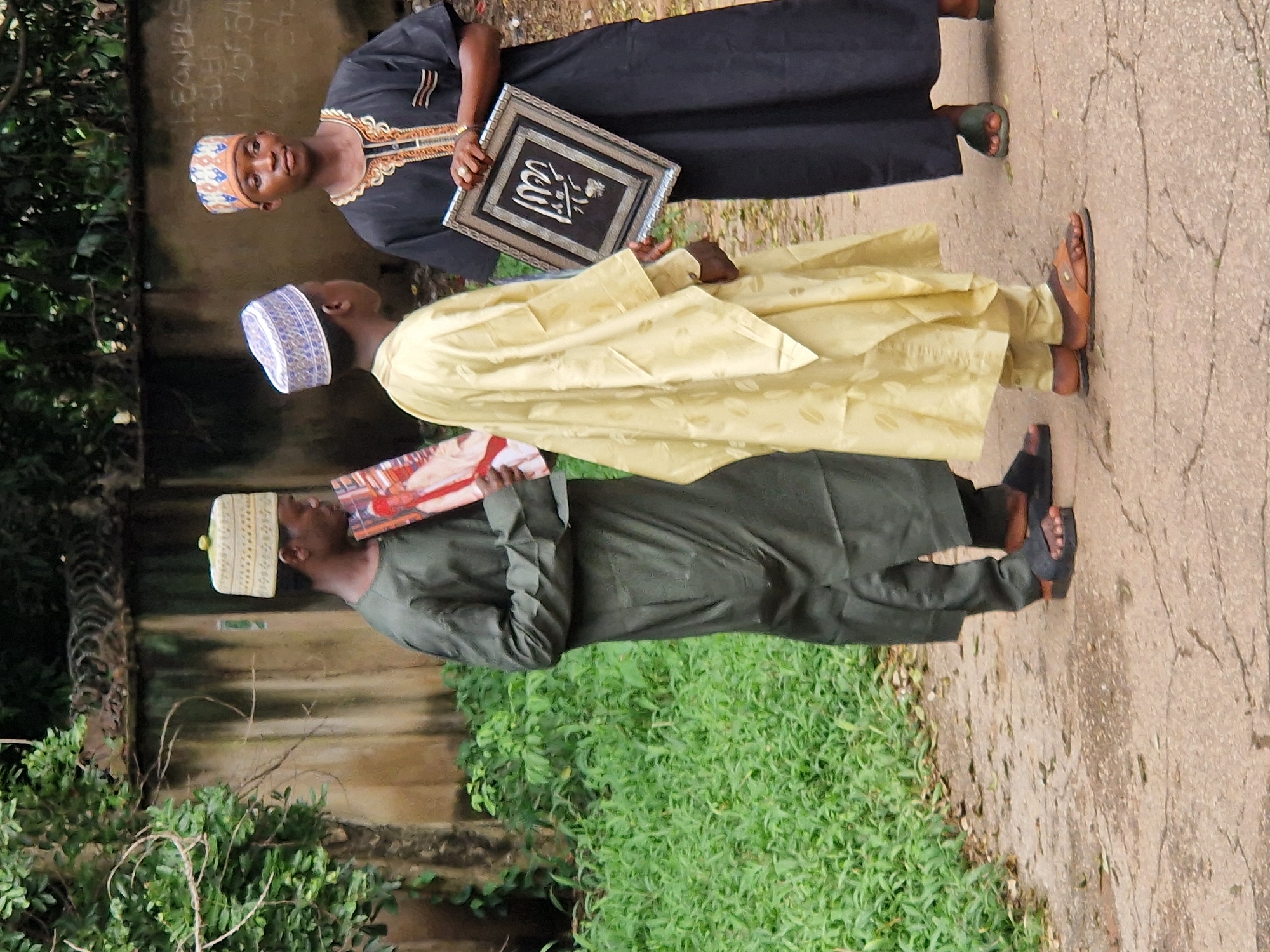 In Jos, Nigeria, three young men stand on a cracked dirt path near a wall overgrown with vegetation. The man on the left wears a dark green robe and a white cap, holding a red-covered book. The man in the middle is dressed in a light yellow robe and a blue and white patterned cap. The man on the right wears a black robe with gold trim and a patterned cap, holding a framed piece of art with Arabic calligraphy. The environment suggests an outdoor, possibly informal setting.
