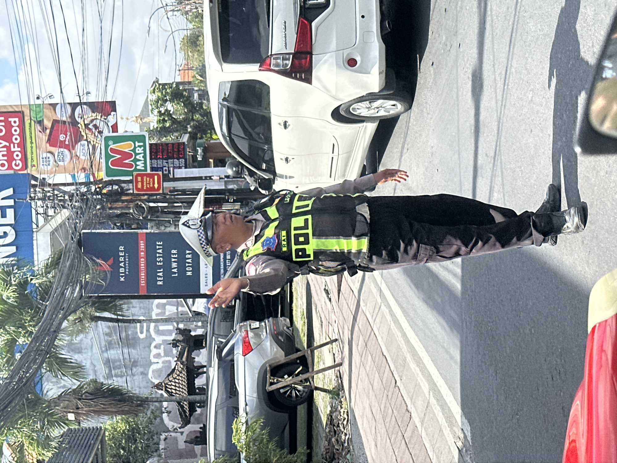 A traffic policeman wearing a reflective vest with "POLISI" written on it is directing traffic on a street in North Kuta, Indonesia. He is wearing a white and blue cap and dark trousers. The policeman's right hand is raised in a gesture to stop traffic. In the background, a white minivan and a grey SUV are parked. There are several signs in the background, including one for "MART" and another for "KIBARER PROPERTY". The scene is illuminated by bright sunlight, casting shadows on the road. The overall environment appears to be an urban setting with buildings and trees visible.