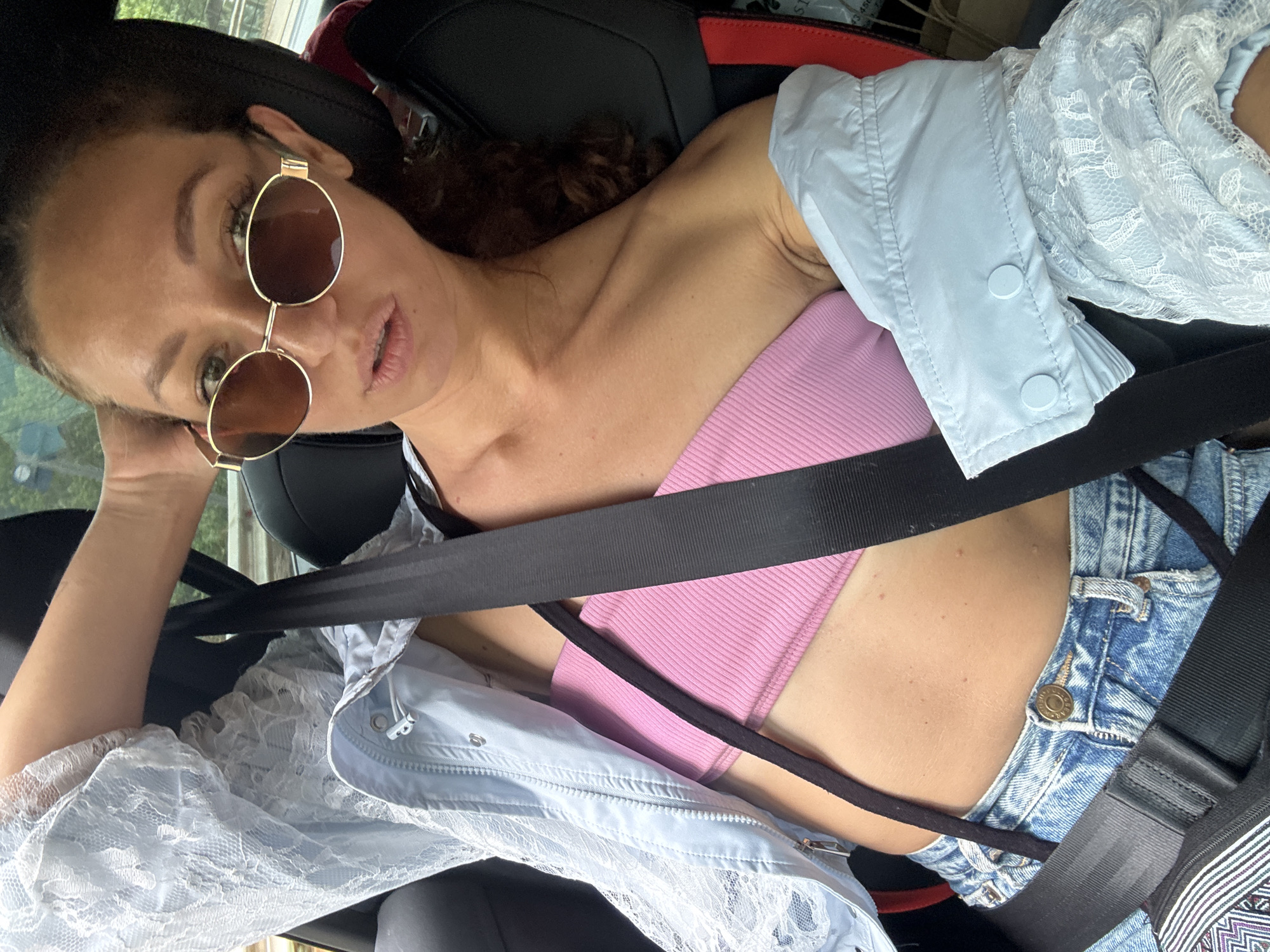In the city of Dế Xu Phình, Vietnam, a female individual is observed seated in the front passenger seat of a vehicle. The individual has dark, curly hair visible and is wearing oval-shaped sunglasses with gold-colored frames. Her lips are slightly parted. She is dressed in a pink ribbed strapless top, partially covered by an open, light blue jacket featuring intricate white lace on the sleeves and body. The jacket displays a zipper and white drawstring toggles on the left side, and two light blue buttons on the cuff of the right sleeve. Distressed blue denim jeans with a gold-toned button are visible at her waist. A black seatbelt is securely fastened diagonally across her chest and abdomen, with the buckle mechanism visible on her left hip. A thin black strap is visible across her midriff. Her right arm is raised, with her hand resting on the back of her head. The vehicle's interior features black seats with minor red accents on the seat back. Through the window, blurred green foliage and a roadside barrier are visible in the background.