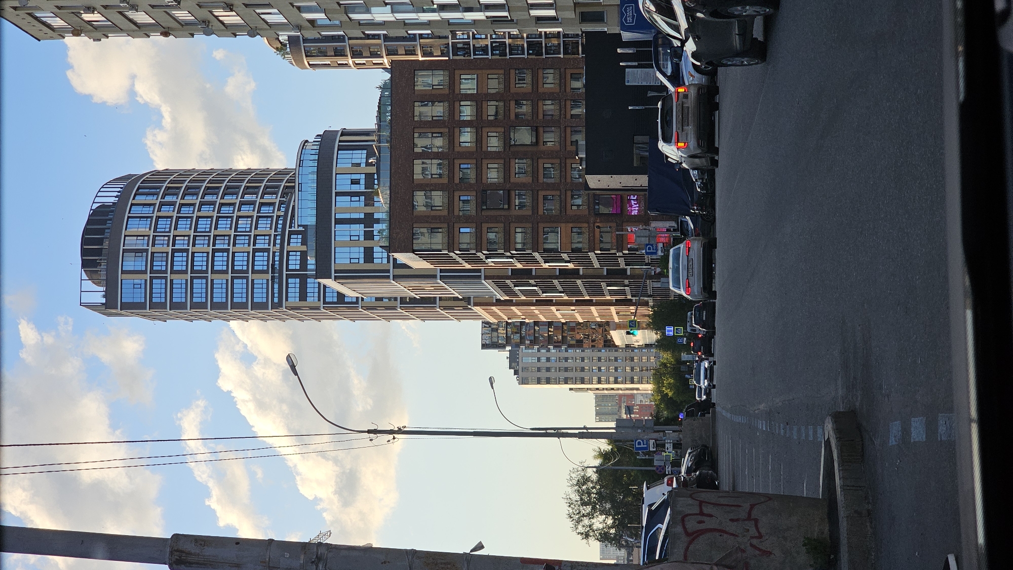 This is a vertical, eye-level, full shot image of a street scene in Yekaterinburg, Russia, on a clear day with scattered clouds. The foreground features a dark asphalt road with white dashed lines and a concrete curb on the left side, marked with red graffiti. Several cars are visible on the road, with some parked on the left and others driving. In the background, a row of modern, multi-story buildings dominates the skyline.
The most prominent building is a tall, curved tower with a grid of blue windows and lighter-colored vertical accents, topped by a dark rooftop structure. Adjacent to it are other contemporary buildings, some clad in brick and others with darker facades and windows. A street lamp with a curved pole extends into the frame from the left. Traffic signs, including parking indicators, are visible near the sidewalk. The sky is a clear blue with soft, white clouds. The overall impression is of a developing urban area with modern architecture.