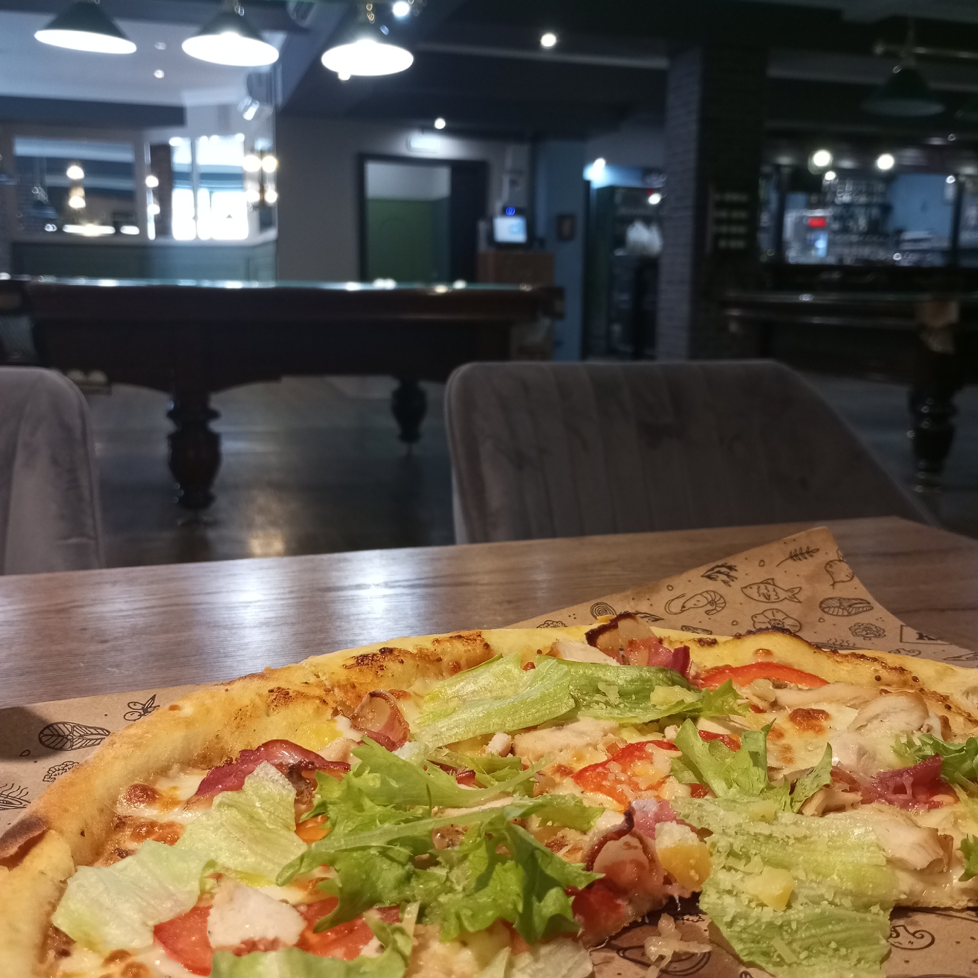 An overhead, close-up shot shows a slice of Caesar salad pizza on a wooden table in a restaurant. The pizza is topped with lettuce, tomatoes, bacon, and chicken. In the background, a pool table and bar area are visible. The restaurant has a modern industrial design with exposed brick and metal accents. The lighting is dim and moody, with spotlights illuminating the bar and pool table. The image is slightly blurry, suggesting it was taken in low light conditions. The restaurant is likely located in Izhevsk, Russia, as indicated by the prompt.