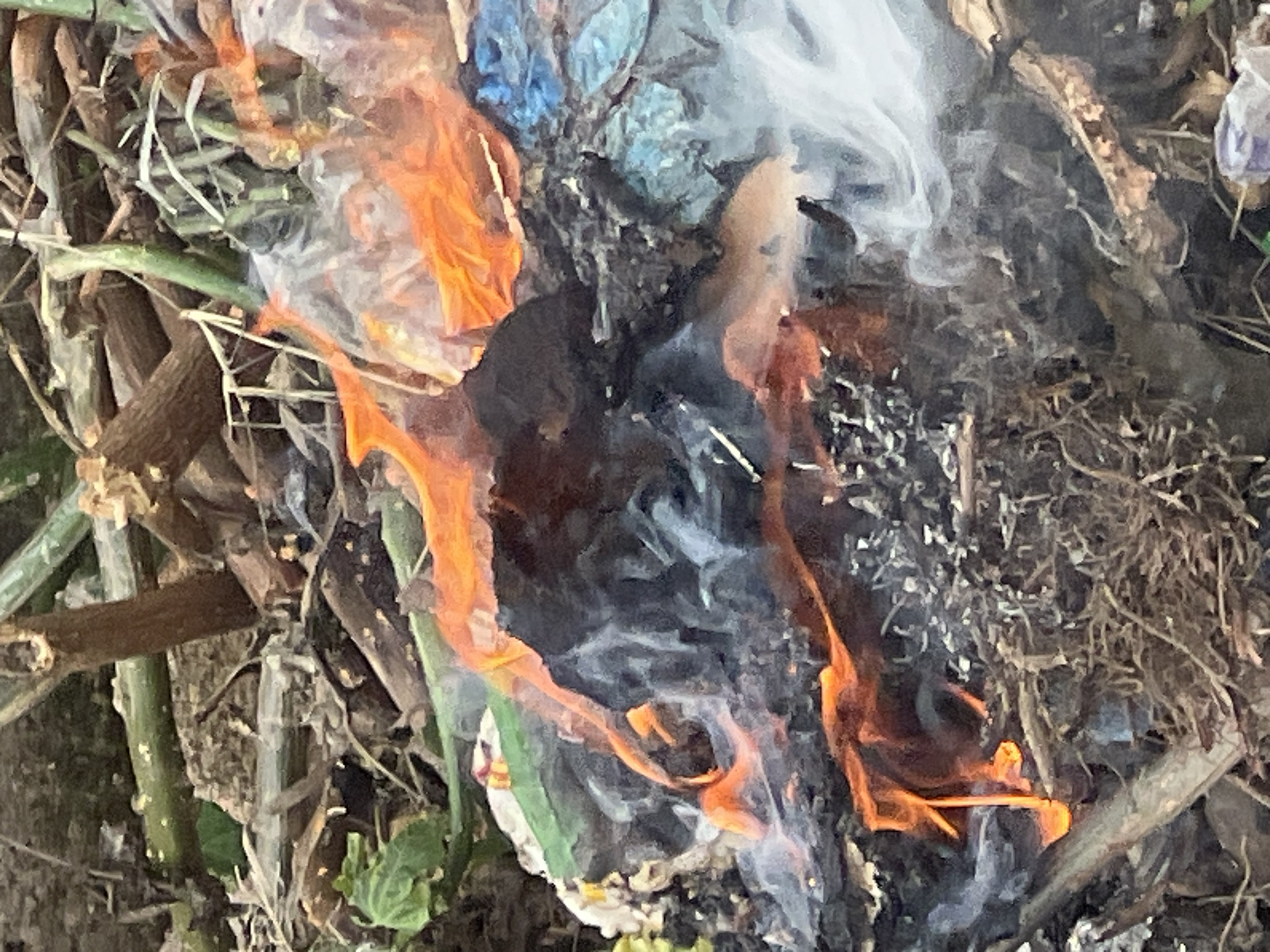 The image depicts a fire burning debris in Gura Top, Nigeria. The fire is consuming various materials, including plastic, vegetation, and other unidentified objects. White smoke billows from the burning debris. Some green plant life is visible around the edges of the burning pile.