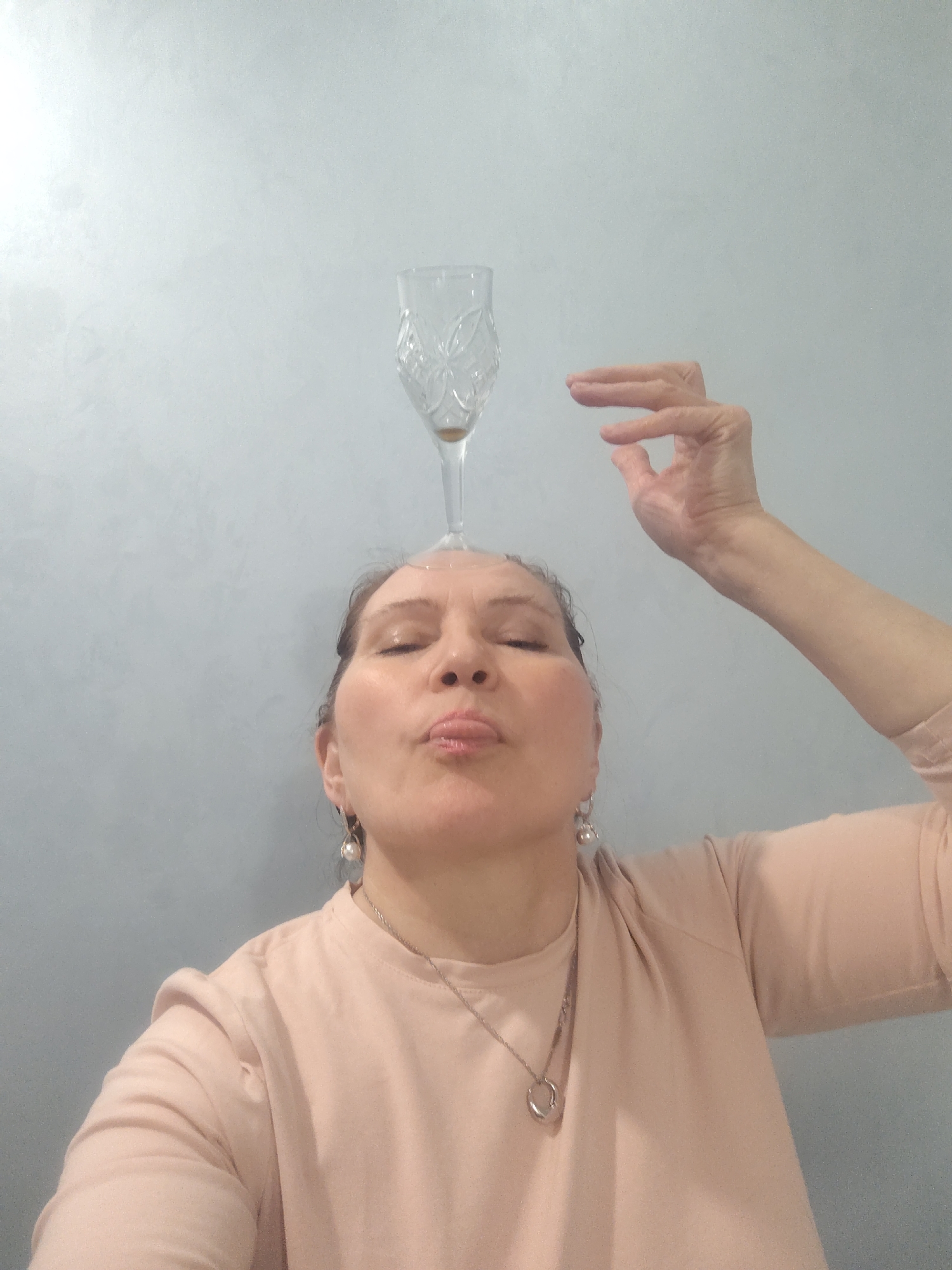 A woman balances a champagne flute on her head, appearing to maintain stability by positioning her hand nearby. She wears earrings and a necklace, and her face is puckered in concentration. The background is a plain wall. The image provides no indication of a specific location or association with Sarapul, Russia.
