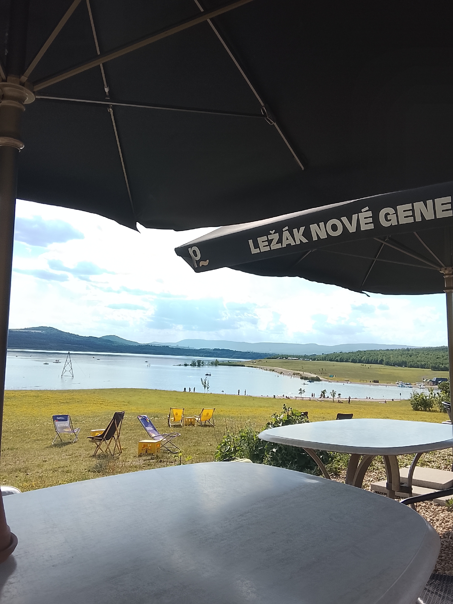 The image depicts an outdoor seating area of a establishment with a view of a large body of water, likely a lake, and a distant landscape of rolling hills and forests under a cloudy sky. In the foreground, two tables are visible, one closer and larger, and another further away. The tables are a light gray color with a smooth surface. To the left and right of the tables are outdoor umbrellas, one dark and larger, covering a portion of the foreground.
Beyond the seating area, a grassy field dotted with yellow flowers stretches towards the water. Several beach chairs are scattered across this field, including yellow loungers and some with blue and white striped fabric, one of which has the word "BARELL" printed on it. Some chairs appear to be made of plastic crates.
The body of water is calm, with several small boats and people visible near the shore. In the middle of the lake, a tall metal structure resembling a transmission tower or a ski jump support is present. The far shore of the lake is lined with trees and more rolling hills, with a hint of a town or industrial area in the far distance. The sky is bright with scattered clouds, suggesting it is daytime. The overall scene conveys a relaxed, possibly recreational atmosphere at a waterfront venue.