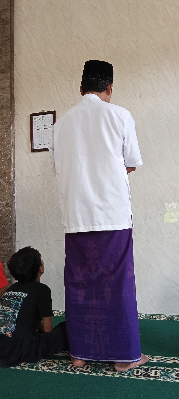 The image shows two men standing in a room with beige tiled walls. The man in the foreground is wearing a white shirt and a purple sarong. He is wearing a black skullcap. The man in the background is partially visible and is wearing a black shirt. There is a framed document on the wall, and the room has a green carpet on the floor. The coordinates provided, -6.973 latitude and 107.521 longitude, place the scene in a mosque in Kecamatan Kutawaringin, Indonesia.