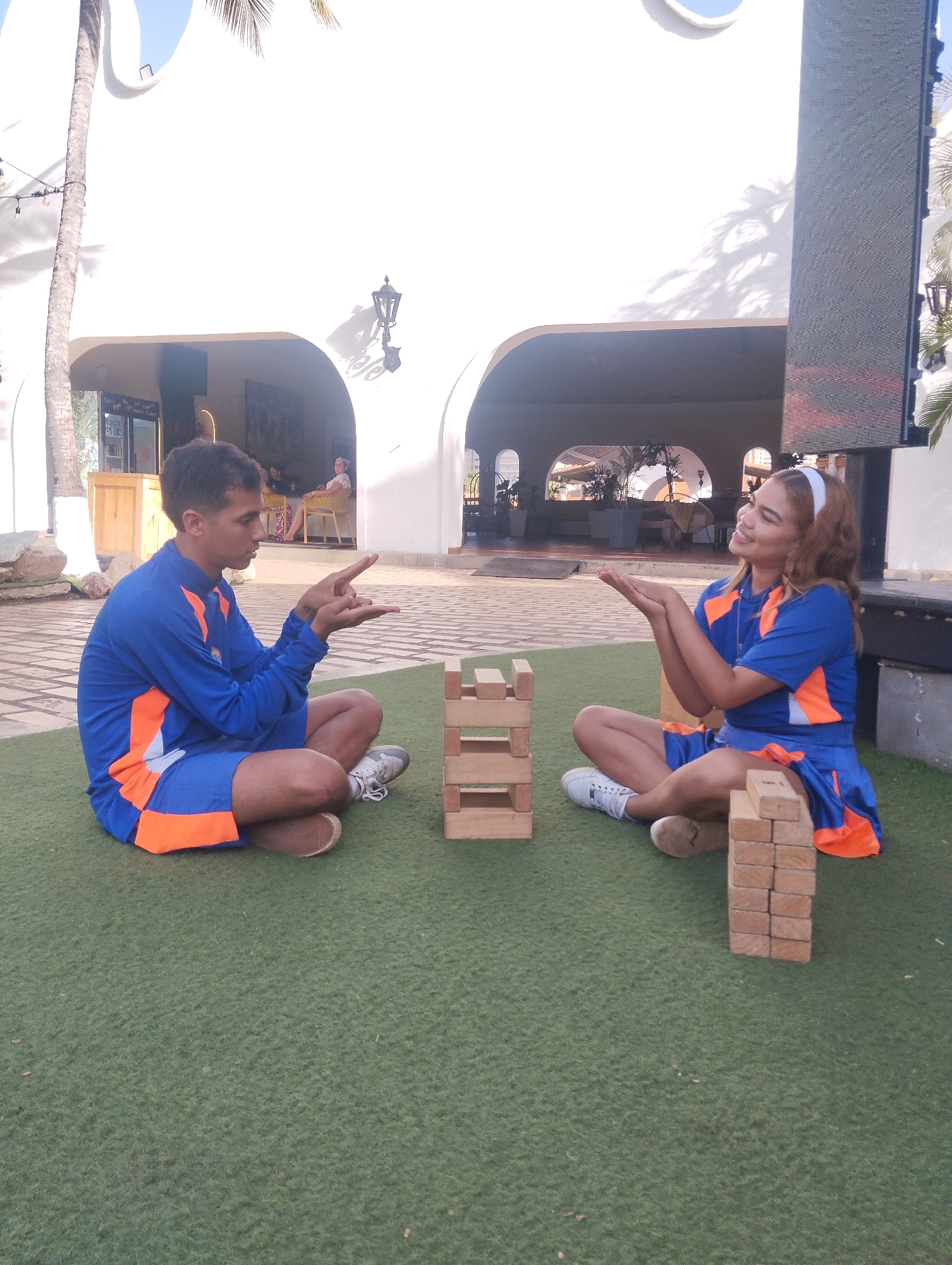 Two individuals, a male and a female, are sitting cross-legged on a green artificial turf. They are both wearing matching outfits consisting of a blue long-sleeved shirt with orange accents and blue shorts also with orange accents. Between them, a wooden block tower is constructed. The male is on the left, and the female is on the right. Both are gesturing with their hands towards the tower. The male is darker-skinned, while the female is lighter-skinned and wearing a white headband. Behind them is a building, possibly a hotel or resort, with white arched openings and a lamppost. The photo was taken in Porlamar, Venezuela.