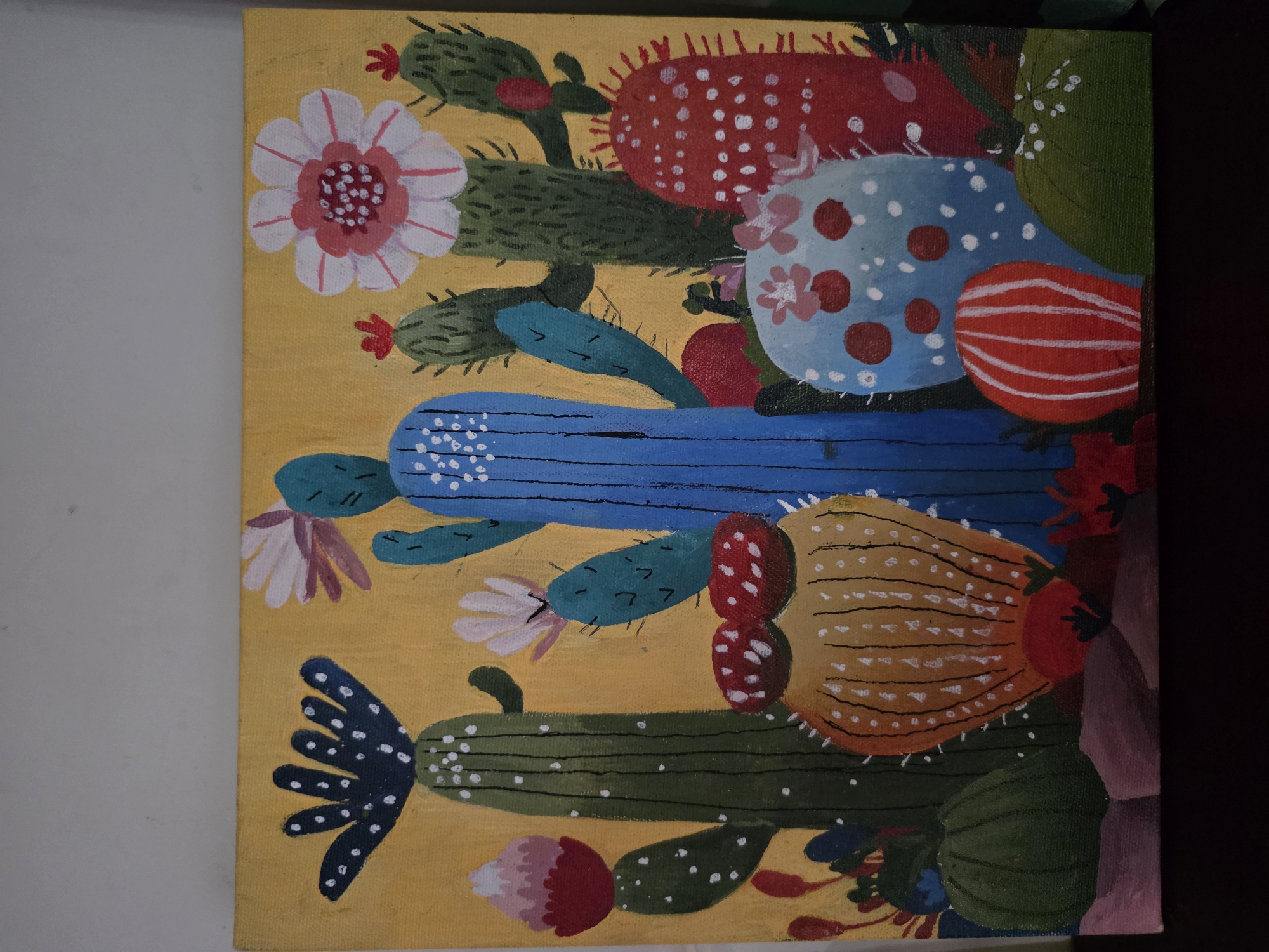 This is a colorful, stylized acrylic painting of various cacti and desert plants on a yellow background. The painting features several tall, green cacti, one prominent blue cactus, and a red barrel cactus. There are also smaller, rounder cacti in blue and orange, adorned with white dots and red markings. Several abstract flowers in shades of pink, white, and red bloom among the cacti. The foreground depicts a rocky, purple and brown landscape. The painting is framed by a dark surface, likely a table, at the bottom. The background wall is a plain, light gray. The image does not contain any specific landmarks or elements that would allow for the determination of its location in North Kuta, Indonesia.