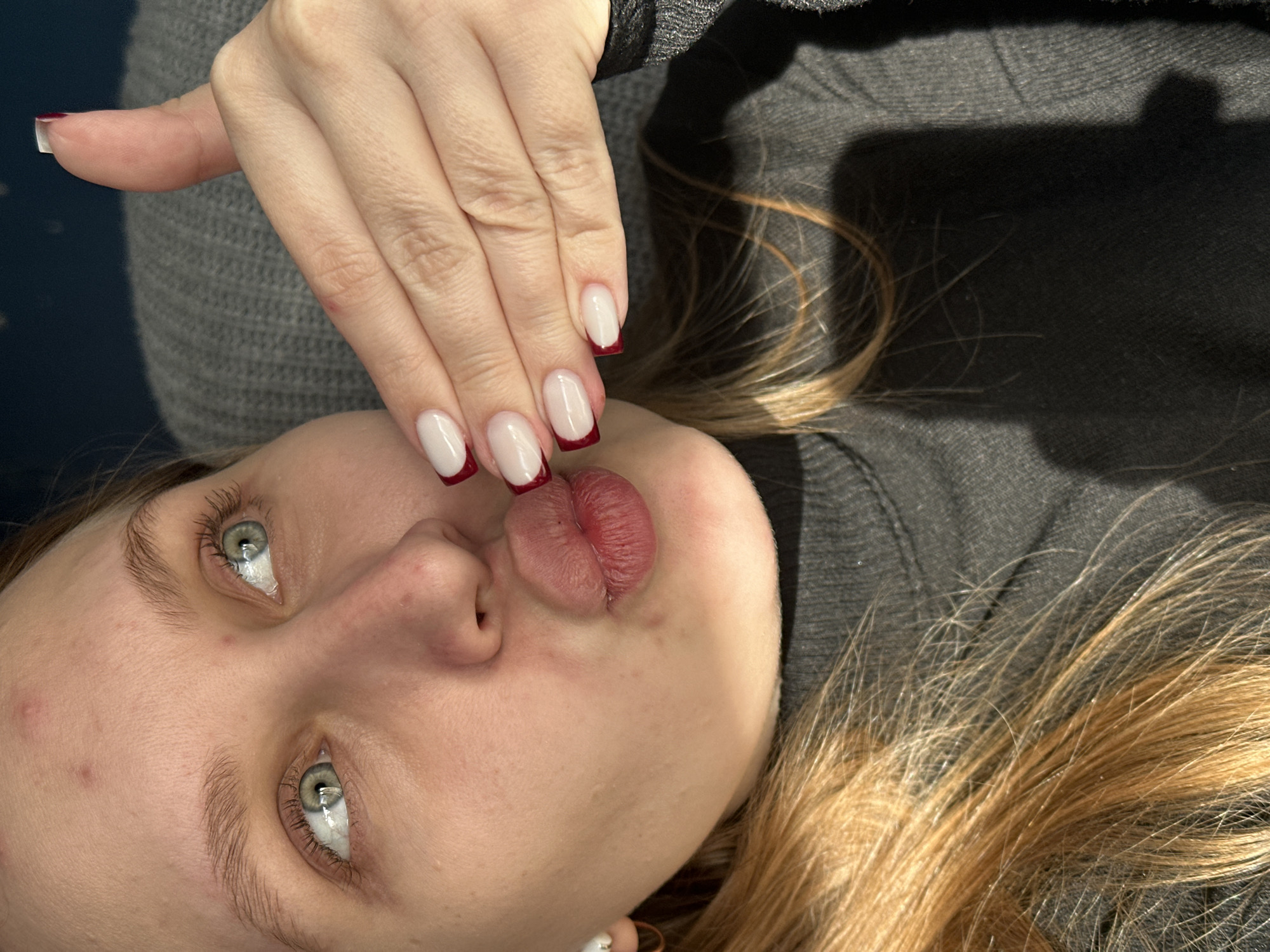 This close-up, high-angle portrait features a young woman, likely taken indoors in Chaikovsky, Russia. She is positioned as if lying down, looking directly upwards towards the camera, with a playful expression.
The central subjects are the woman's face and her left hand. Her fair skin, striking light green or hazel eyes, and light brown eyebrows are prominent. Her lips are dramatically puckered into a "kissing" or "fish face" pose, showing their natural reddish tone. There are minor blemishes and pores visible on her skin, reflecting a natural appearance. Long, light brown or blonde hair cascades around her head, resting against a dark grey background, possibly a garment.
Her left hand is held above her face, showcasing a set of well-maintained nails. The nails feature a fashionable French manicure, characterized by a clear or pale pink base coat and distinct, neatly applied dark red or burgundy tips. The nails are of medium length and have a squared-off shape. She is wearing a textured grey sweater, visible around her shoulder and upper arm, which blends into the dark grey or black fabric that forms the background.
The lighting is bright and direct, creating reflections in her eyes and highlighting the details of her nails and skin. There is no visible text within the image. The overall impression is a casual and posed shot, emphasizing the woman's features and her styled nails.