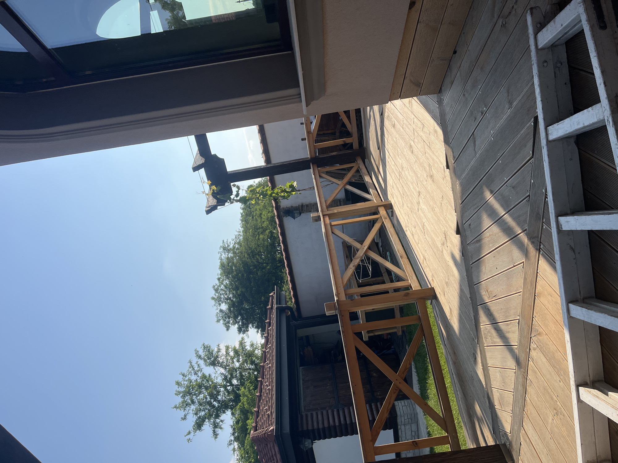 The image depicts an outdoor residential or recreational area in Войводино, Bulgaria, under a clear, bright blue sky. A wooden deck, constructed from light-colored planks, occupies the foreground and extends into the midground, exhibiting distinct shadows cast by strong sunlight, indicative of a sunny day. In the immediate foreground, a white ladder rests partially visible on the deck. A wooden railing, featuring X-shaped cross-bracing, separates the deck from a grassy area beyond.
To the left, beyond the railing, stands a single-story building with a dark, tiled, pitched roof and a log-cabin style lower section, featuring a dark wooden door and an exterior light fixture. Behind this structure and extending to the right is a light-colored wall. A dark wooden pergola, draped with green vines, is positioned in the midground behind the railing, in front of the white wall. A wooden cart wheel is partially visible behind the railing near the pergola and wall.
On the right side of the image, a section of a building, possibly a house, is visible, featuring a light-colored, textured facade with dark-colored window trim around a large window. The window reflects the clear sky and distant greenery. Below the window, there is a decorative molding and a lower section clad with horizontal wooden planks. In the background, numerous green trees rise above the structures. No individuals are present in the scene.