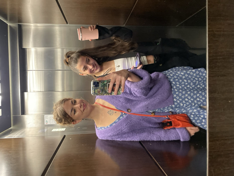 Two young women are seen in a mirror selfie taken inside an elevator. The image is rotated 90 degrees counter-clockwise, making the subjects appear on their sides.
The woman closer to the camera is on the left side of the frame (when upright). She has light brown hair, styled in soft waves around her face, and is smiling towards the camera. She wears a fuzzy, light purple open-knit cardigan over a blue dress with a small white floral pattern. A small red cross-body bag hangs from her shoulder. Notable details include sparkling makeup near her eyes and a small tattoo on her upper chest/shoulder area. She is holding a smartphone with a decorated case to take the picture, which is reflected in the elevator mirror.
The second woman is positioned behind and slightly above the first (when upright). She has darker brown hair pulled up in a half-ponytail and is also smiling broadly. She is dressed in a black jacket over a white top and is holding a pink thermal mug in her hand.
The setting is an elevator with reflective, polished metal walls, likely stainless steel. These walls show reflections of the women and the opposite side of the elevator. There are also darker, possibly wood-paneled, sections visible at the top and bottom edges of the frame.
The activity is a casual mirror selfie, capturing a moment of apparent happiness and friendship between the two women. Both are smiling, conveying positive emotions.
On the elevator wall, in the background, there are some small, rectangular signs or labels, one of which appears to have a red border. The text on these signs is too small and indistinct to be legible due to reflections and image resolution. The location context for this event is Boreti, Montenegro.