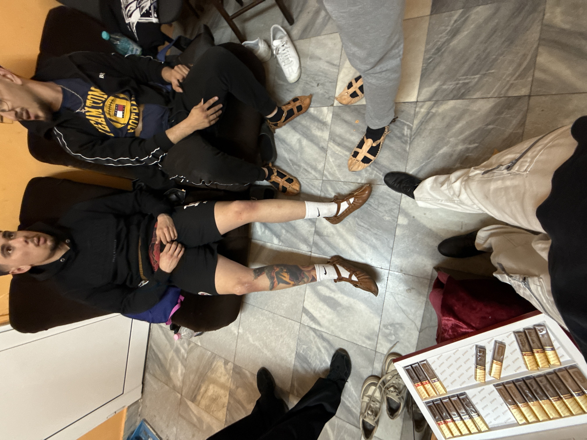 Four individuals are in a room with a marble-tiled floor in Buhovo, Bulgaria. Two sit on orange chairs, one wears a black hoodie and shorts, the other a black jacket and blue shirt. They are wearing white socks with brown leather shoes of an ancient roman style. A third person stands in gray sweatpants and wears the same roman shoes. The fourth person, partially visible, wears light pants and black dress shoes. A box of chocolate cigars sits on the floor, along with pairs of sneakers and dark shoes.