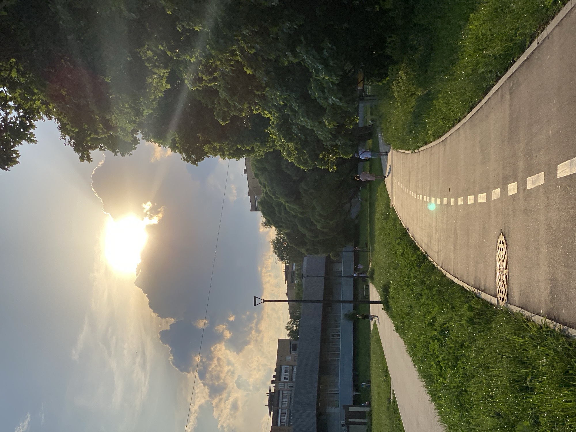 The image captures an urban park scene at sunset. A curved asphalt path, marked with dashed white lines, winds through the frame. Dense green trees and foliage border one side of the path, while a grassy area with occasional benches and people sits on the other. In the distance, residential buildings are visible. The sun, partially obscured by clouds, dominates the sky, casting a bright, warm light across the scene. A lamppost stands between the path and the buildings. A manhole cover is seen on the path. Two figures are walking along the sidewalk. Several people are sitting on a bench.