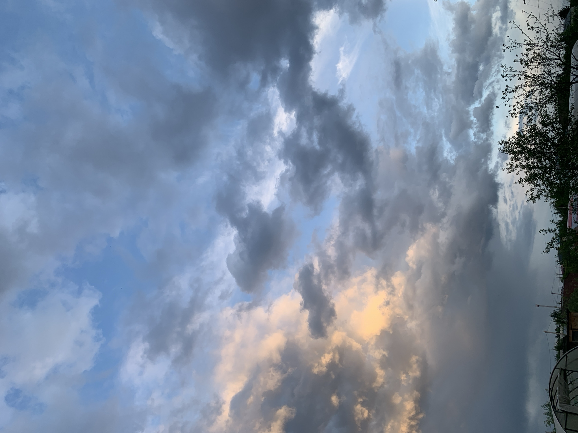 The image captures a sky filled with dark, ominous storm clouds over the city of Chaikovsky, Russia. The clouds are a mix of dark gray and patches of lighter, almost golden-tinged clouds, possibly reflecting sunlight. The sky is only visible in small sections, with the main part being dominated by the clouds. A small amount of green foliage of trees is present on the right edge of the frame. There are objects from the city, like a greenhouse and other unknown buildings and objects. The image is taken from a low angle, looking upwards.