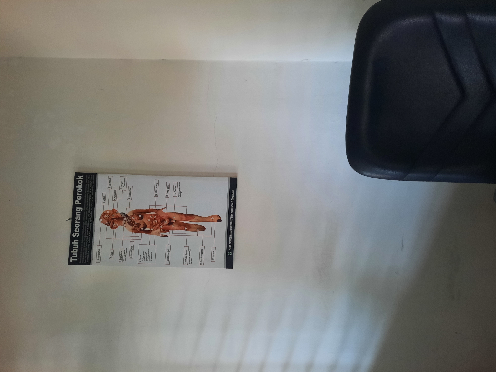 The image depicts a poster titled "Tubuh Seorang Perokok" (The Body of a Smoker) hanging on a lightly textured, off-white wall. The poster is a diagram illustrating various negative health effects of smoking on the human body, with numbered callouts pointing to different anatomical areas and conditions. A dark, upholstered office chair is partially visible in the foreground, its backrest occupying the lower right portion of the frame. The lighting suggests an indoor environment, possibly a waiting area or office. The poster is dated 2006 and attributed to "Pusat Promosi Kesehatan Departemen Kesehatan RI" (Health Promotion Center, Ministry of Health of the Republic of Indonesia), indicating it's an official public health awareness material. The overall impression is one of a healthcare facility or educational setting within the city of Lhokseumawe, Indonesia.
