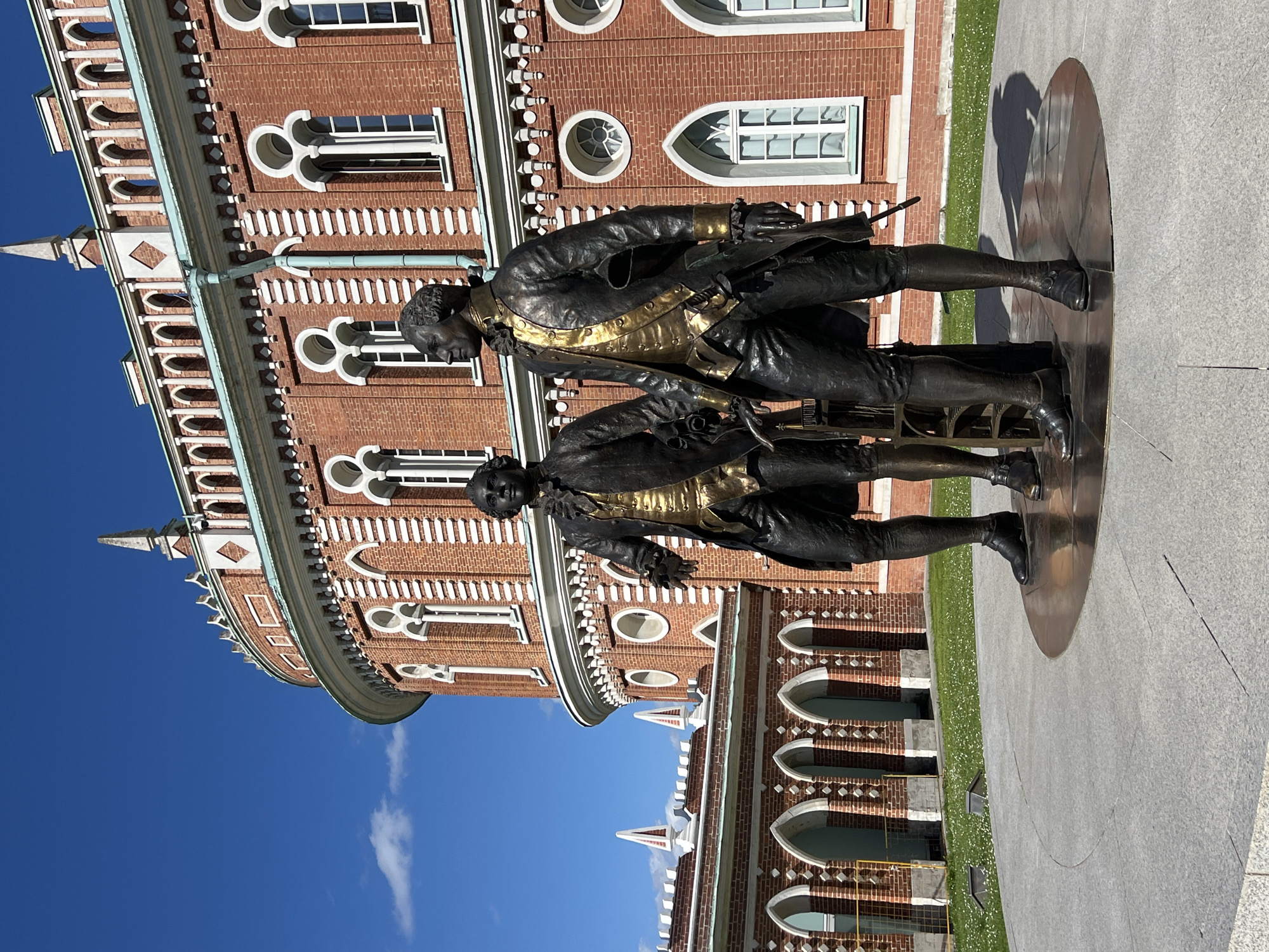 The image shows two bronze statues of men in historical attire standing on a circular base. The statues are positioned in front of a large brick building with arched windows and decorative white trim. The building is curved and appears to be part of a larger complex. To the left of the building, there is an arched walkway with more brickwork. The scene is set outdoors under a clear blue sky with a few wispy clouds. The ground in front of the statues is paved with light grey stone, and a patch of green grass with small white flowers is visible behind the statues. This scene is located in Tsaritsyno Park in Moscow, Russia.