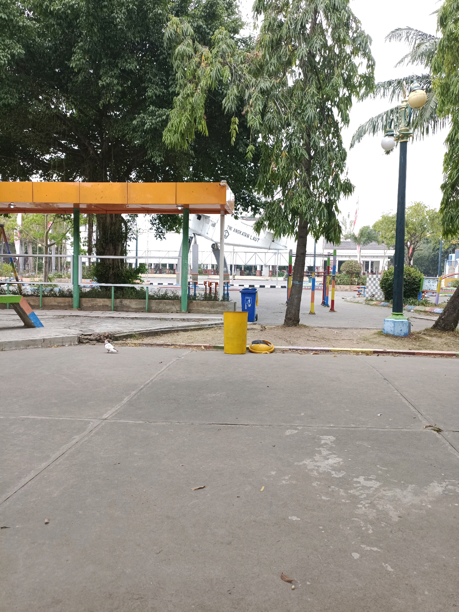The image depicts an outdoor scene in the city of Lamongan, Indonesia, likely a public area such as a park or playground. In the foreground, a gray concrete surface is visible, marked with expansion joints. To the left, a yellow and orange covered structure with green supports is present, possibly a bus stop or shelter. A broom with a blue handle and brush lies on the ground near the shelter. Further back, several brightly colored poles, likely part of a playground apparatus, stand on a dirt and gravel surface. In the middle ground, a white dove is standing on the concrete. Two trash bins, one blue and one yellow, are situated to the left of the colored poles. Behind these, a white building with a red roof is visible, alongside trees and a lamppost with two globe-shaped lights. The background features more trees and a large white structure with text that reads "TNI ANGKATAN LAUT," indicating a naval connection. The overall environment appears to be a public space designed for recreation and gathering.