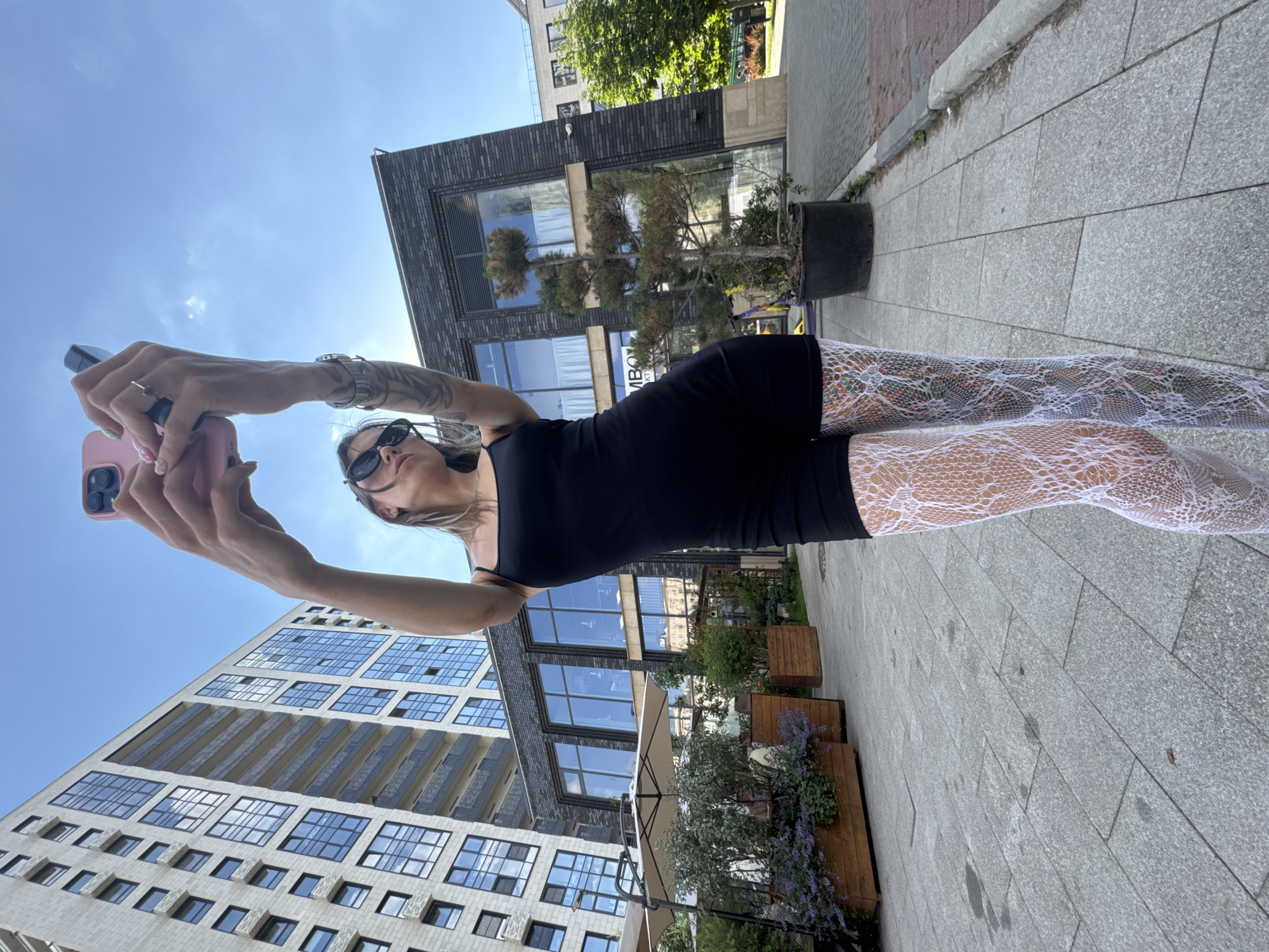 A woman stands outdoors, holding a pink smartphone to take a selfie. She is wearing a black dress, white lace tights, and sunglasses. The scene is urban, with modern buildings and a paved walkway. In the background, there is a multi-story building with a glass facade, a smaller building with dark brickwork, and some greenery in planters. The sky is clear and blue, suggesting it is daytime and the weather is fair. The woman's pose and the selfie stick-like extension of her arm indicate she is actively capturing the moment. The image is oriented vertically, with the sky at the top and the ground at the bottom, and the subjects are arranged horizontally across the frame. There is no visible text in the image.