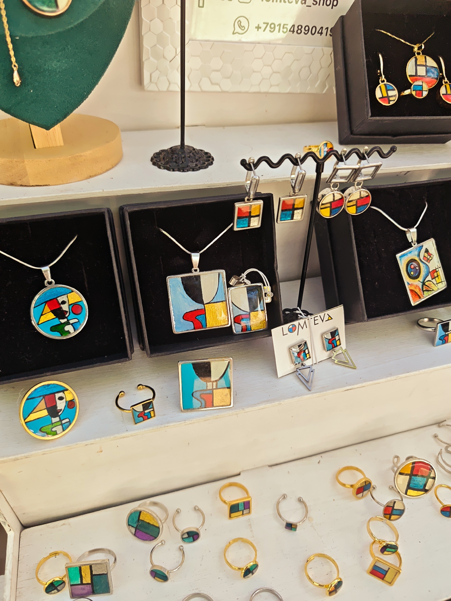 This is a close-up, eye-level, outdoor shot of a display of jewelry, likely at a market or shop, featuring abstract, colorful designs in a style reminiscent of Piet Mondrian. The jewelry is arranged on white shelves and black display boxes.
In the upper left, a green velvet bust displays a gold chain necklace with a circular pendant featuring a colorful abstract design. To its right, a black stand with an ornate base holds a display of earrings.
The main focus is on the jewelry. Several black boxes contain necklaces and matching earrings. The necklaces are silver chains with pendants in circular and square shapes, adorned with geometric patterns of primary colors (red, blue, yellow) and black and white. Matching earrings, also featuring similar geometric designs, are displayed alongside.
Further to the right, on a white shelf, a rack holds several pairs of silver earrings, including dangly geometric shapes and studs with colorful abstract inserts. A small white card with the text "LOMTEVA" and a drawing of earrings is also visible.
The bottom of the image shows a wider assortment of rings, some gold and some silver. These rings also feature small, colorful geometric designs, mirroring the style of the necklaces and earrings.
The background is slightly out of focus but appears to be a wall with a textured pattern and some signage, including a phone number with a Russian country code (+7). Given the presence of Russian signage and the mention of Kimry, Russia in the prompt, this scene is located in Kimry. The overall environment suggests a retail setting for artisanal jewelry. No people are visible in the frame.