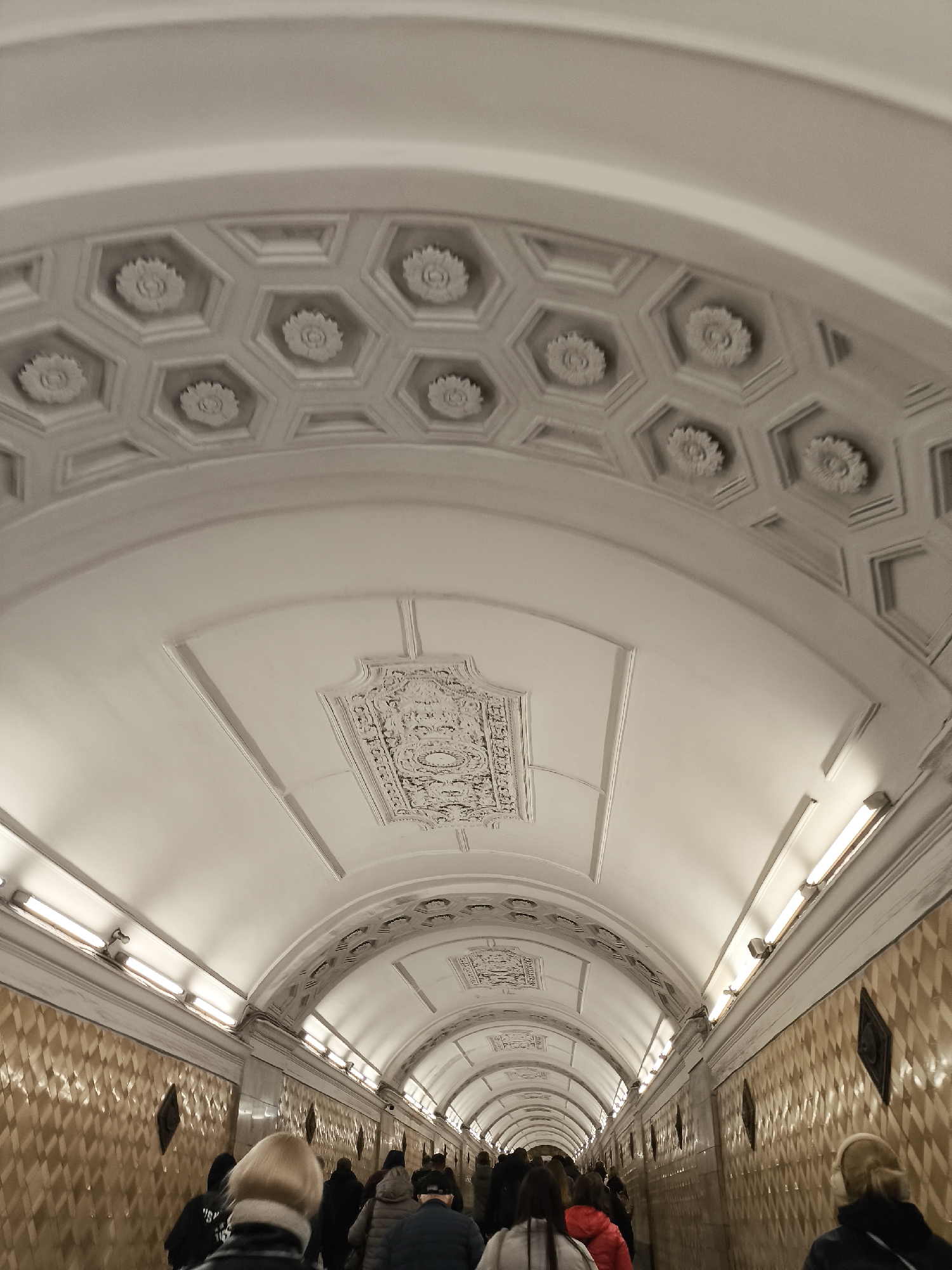 The image captures a perspective view of a long, ornate pedestrian tunnel, likely within a subway or metro system, filled with people walking.
**Subjects:** The primary subjects are numerous people, mostly seen from behind, walking away from the viewer down the length of the tunnel. They are dressed in casual, layered clothing, including jackets and sweaters, suggesting cooler weather. The tunnel itself is a prominent subject, characterized by its distinctive architectural elements.
**Activity or event taking place:** The people are engaged in the act of walking or commuting through the tunnel, indicating a common daily activity in an urban transit environment.
**Notable details:**
The tunnel features a grand, arched ceiling decorated with intricate plasterwork. The section closest to the viewer displays a pattern of hexagonal coffers, each containing a detailed floral rosette in its center. Further down the tunnel, other sections of the ceiling feature different ornate rectangular or shield-shaped decorative panels with elaborate relief patterns.
The walls of the tunnel are clad in a mosaic of light brown and off-white tiles arranged in a diamond pattern. Interspersed along the walls are darker, square or diamond-shaped plaques or decorative elements. The tunnel is illuminated by continuous rows of fluorescent tube lights mounted along the upper edges of the walls, casting a bright, even light throughout the passage. The perspective makes the tunnel appear to narrow and converge in the distance, emphasizing its length. The artificial lighting and interior setting mean the time of day and external weather are not discernible, though the clothing suggests a non-summer season.
**Visible text:** There is no discernible text visible anywhere in the image.