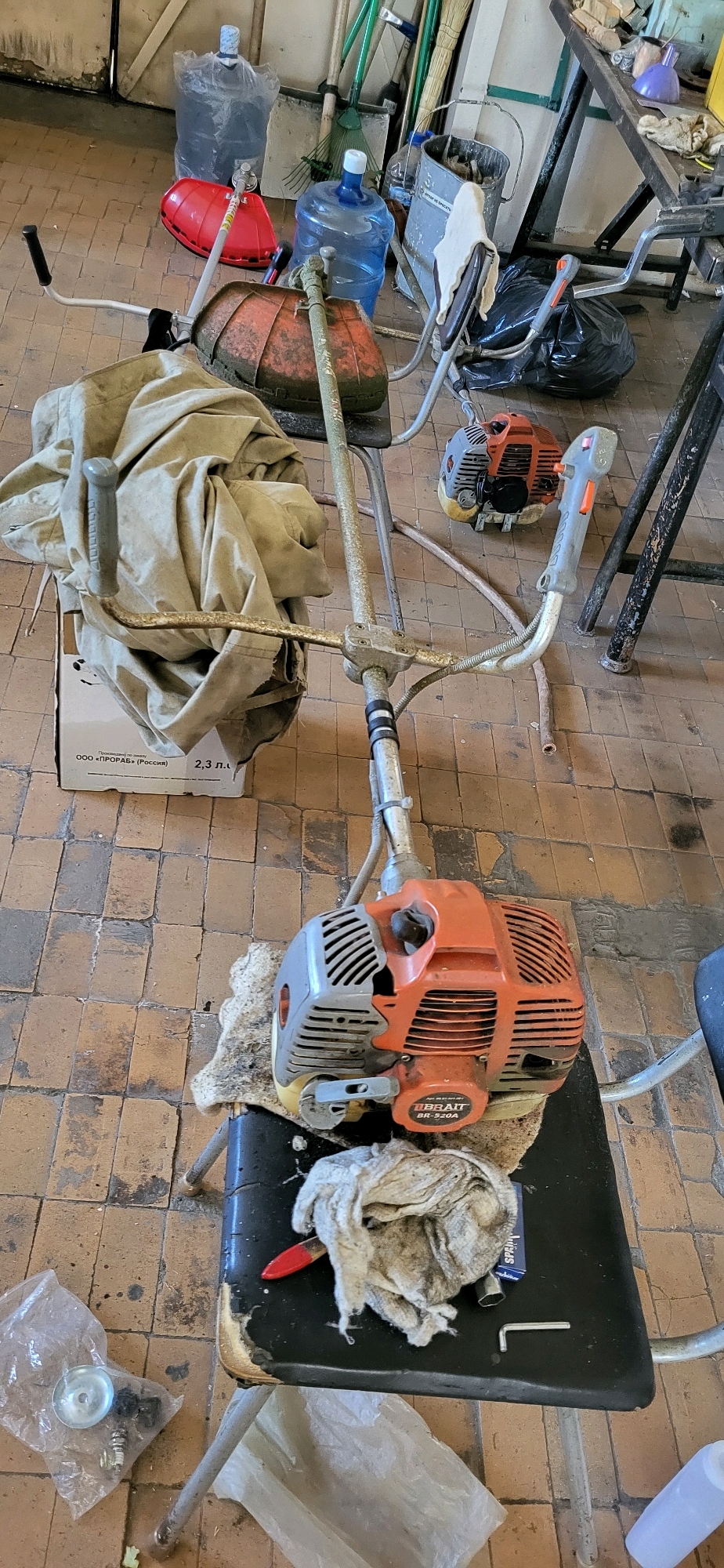 The image depicts a cluttered garage or workshop interior in Oskemen, Kazakhstan. The scene is dominated by various garden tools and maintenance equipment. In the foreground, a disassembled string trimmer lies on a black stool, with its engine unit prominently displayed. The engine is orange and gray, bearing the label "TIBRAIT BR-520A". Various parts of the trimmer, including a fabric cover and a metal handle, are scattered around it.
To the left, a cardboard box with Russian text, possibly indicating product specifications such as "2,3 л.", is partially visible. A clear plastic bag containing metal parts and a silver disc is also present. Nearby, a red attachment, likely a guard for the trimmer, is on the floor.
In the background, several large water bottles, one in a clear plastic bag and another blue one, are situated on the tiled floor. Gardening tools such as rakes and a shovel are leaning against a wall. A metal bucket filled with debris is also visible. A black plastic bag, possibly containing trash or supplies, is placed on the floor behind the stool. The environment appears unkempt, with dust and dirt evident on the floor and objects. The lighting suggests an indoor setting, possibly with ambient light from a window or overhead fixture, casting shadows on the floor. There are no people present in the image.