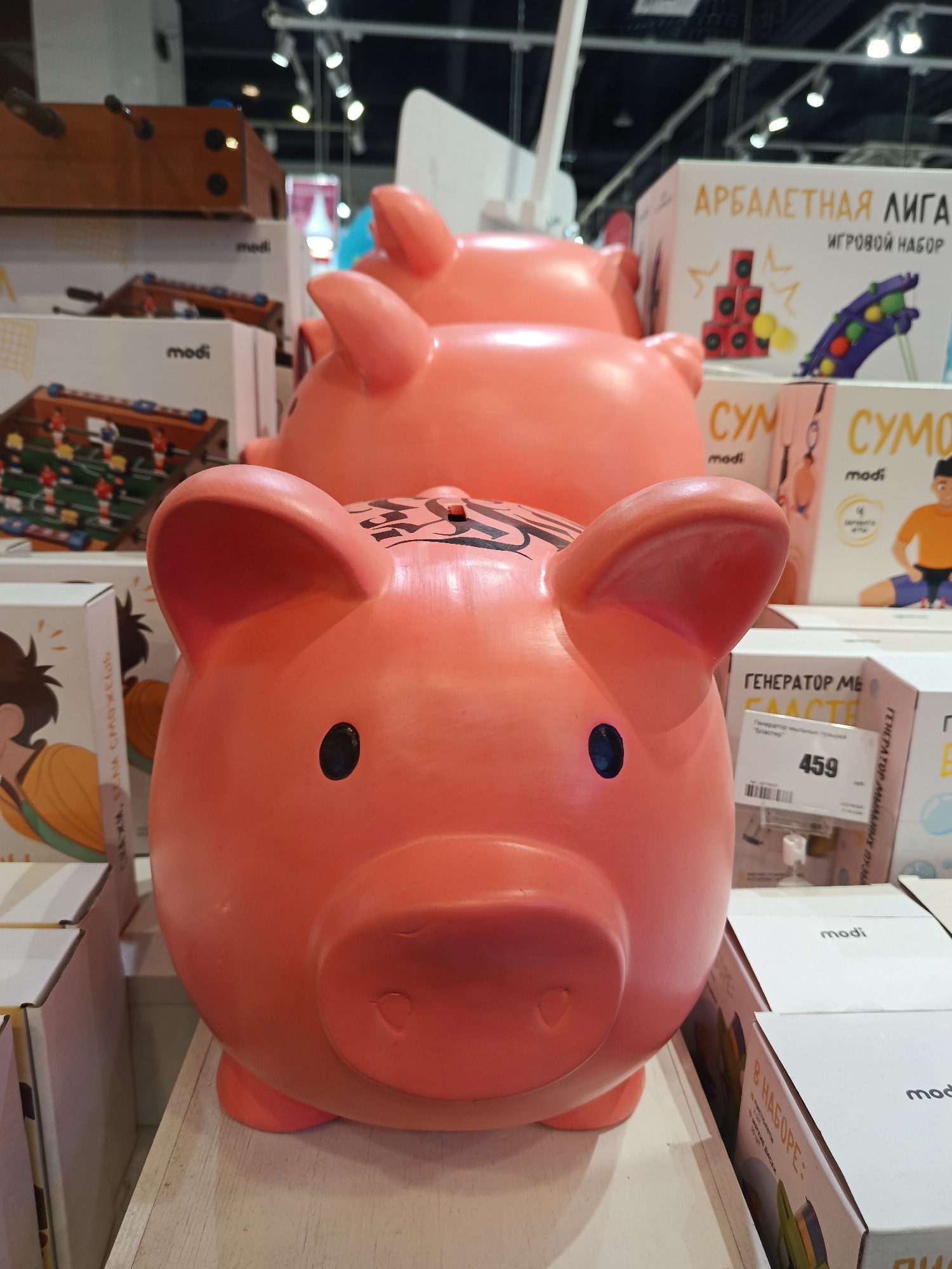 This is an indoor photograph of three bright pink pig-shaped piggy banks arranged in a row, with the closest pig filling much of the frame. The piggy banks are on a shelf amongst other merchandise. Each pig has small, round, black eyes and a prominent snout. The top of the closest pig's head has what appears to be decorative black markings, and a coin slot.
Boxes with the brand name "modi" are visible around the piggy banks. Boxes with text in Russian, like "АРБАЛЕТНАЯ ЛИГА ИГРОВОЙ НАБОР" (Crossbow League Game Set) and "СУМ СУМО" (Sumo Sum) are also seen. A price tag displaying the number "459" is visible. The background shows other toys and merchandise.
The location appears to be a retail store, possibly in the city of Moscow, Russia, as indicated by the Cyrillic writing on the boxes.