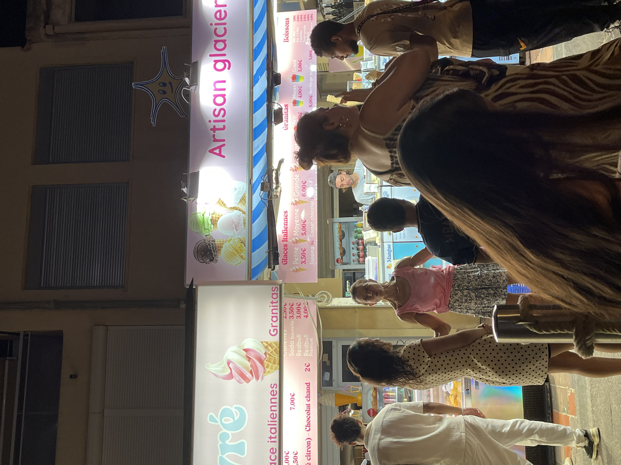 The image depicts an outdoor scene at night, likely a street or promenade, in Palavas-les-Flots, France. The focal point is an ice cream stand with illuminated signage. One sign reads "Artisan glacier" in bright pink lettering. Below this, a pink and white swirl of soft-serve ice cream in a waffle cone is prominently displayed. Another section of signage features text in French, including "Crêpes italiennes" and "Granitas". Prices are listed in Euros, with items like "Cornet chocolat" priced at "2€" and various drinks and ice cream options ranging from "3,00€" to "7,00€". To the right, another sign displays "Glaces Italiennes" with prices for "Petite" and "Moyenne" sizes.
Several people are visible in the foreground and midground, appearing to be customers or staff. A man in a white shirt and trousers stands on the left, facing towards the ice cream stand. A woman in a polka-dot dress is standing next to him, looking towards the stand as well. In the center, a woman wearing a pink tank top and a patterned skirt is speaking. Behind her, a man in a dark shirt is visible looking towards the counter. On the right, a man in a light brown t-shirt and dark pants stands looking at the ice cream display. The environment suggests a commercial area with a lively atmosphere, likely popular with tourists. The lighting is artificial, coming from the signs and overhead fixtures, creating a warm glow on the scene.