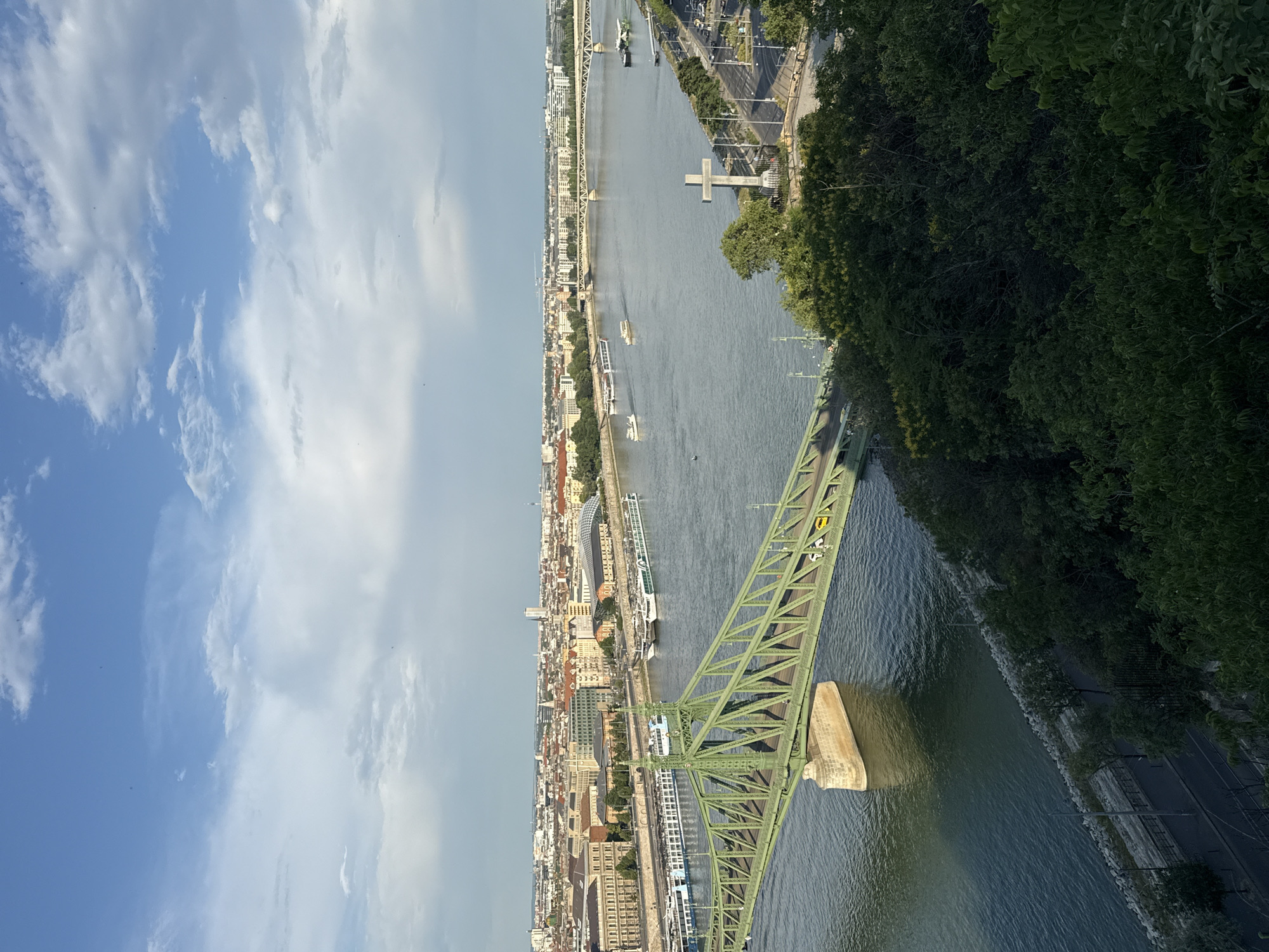 This is a high-angle, wide-shot photograph of the Liberty Bridge in Budapest, Hungary, crossing the Danube River. The bridge, painted green, spans the river with its distinctive truss structure and is supported by stone pillars. Several cruise ships and smaller boats are visible on the water.
Across the river, the city skyline of Budapest is visible with numerous buildings, including some with traditional architecture and others more modern. A prominent white cross monument stands on the Gellért Hill side of the riverbank. The sky is a mix of blue and white clouds, suggesting fair weather. The foreground is dominated by lush green trees and foliage, indicating this viewpoint is from a higher elevation, likely on Gellért Hill. A road with some traffic is visible along the riverbank on the far side of the bridge. The overall scene captures a scenic urban landscape with a focus on the iconic bridge and the natural beauty of the river and surrounding hills.