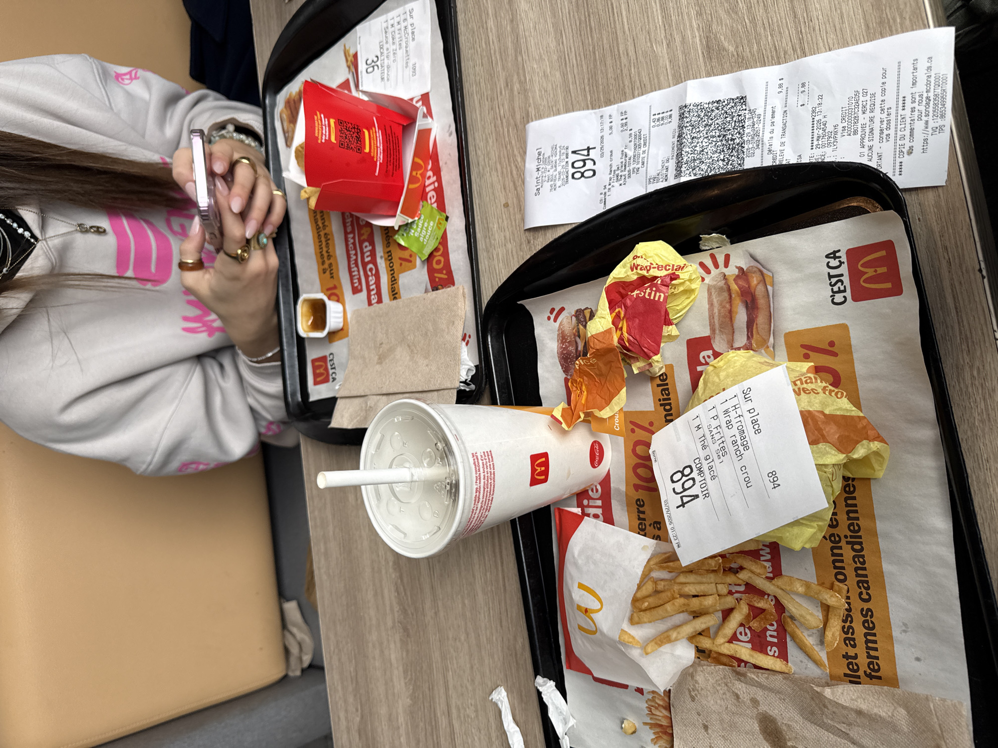 The image shows a McDonald's meal on a table, likely at a location in Montreal, Canada. There are two trays, one with a box of Chicken McNuggets, sauce packets, and a dipping sauce container. The second tray contains the remnants of a meal, including a drink, French fries, burger wrappers, and a receipt. A person is seen in the background holding a phone.