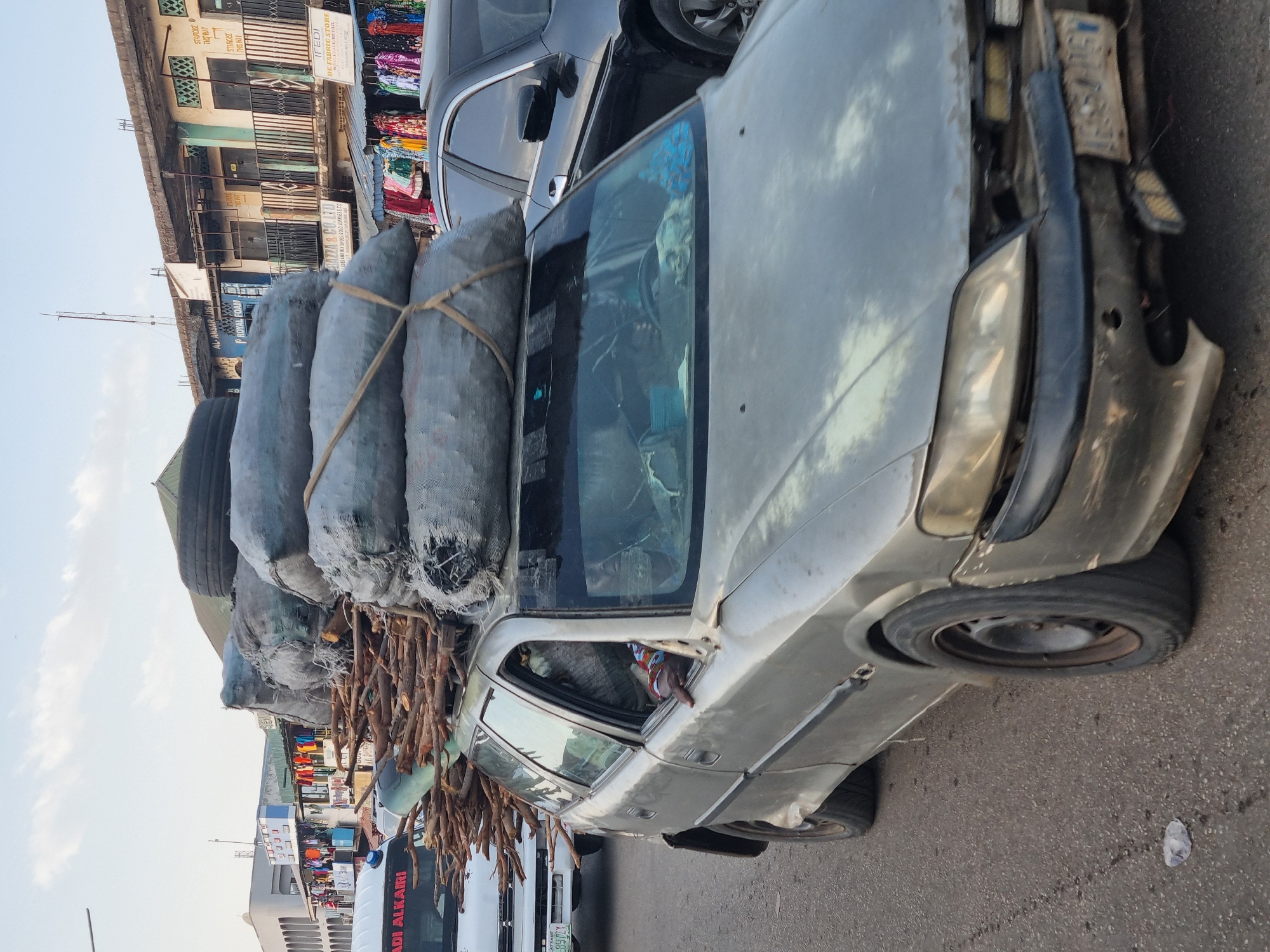 A car is overloaded with bags, charcoal, and wood on a street in Jos, Nigeria. The car, appearing in poor condition, has bags strapped to its roof, obscuring the driver's view. Additional bundles of sticks protrude from the sides. The vehicle is parked on a paved road amidst an urban environment. Buildings, shops, and pedestrians are visible. Nearby is a bus with "ADI ALKAIRI" and "897ZY" printed on it. A sign indicates "AL-ANSAR LOS ROYAL". Another sign reads "STAIRCASE THIS WAY" along with "IFEDI". Clothing items hang for sale. The overall scene shows heavy traffic in a bustling commercial area.