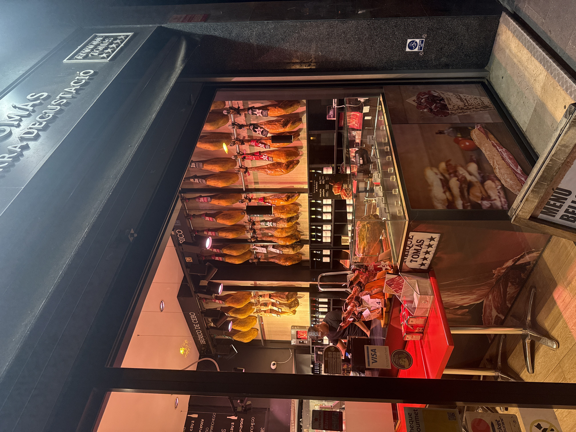 The image shows a nighttime street view of a Enrique Tomás, a gourmet ham shop located in Barcelona, Spain. The shop's facade is brightly lit, with a large display window showcasing numerous hanging cured hams, also known as jamón ibérico. Inside, a person is working behind a counter, possibly a cashier or employee, and a Visa payment sign is visible. The interior also features shelves stocked with wine bottles and more ham products on display. Outside the shop, a red table with a chrome base is set up, and a wooden menu board is placed on the sidewalk. The shop's signage indicates "Enrique Tomás Sierra Degustació," with a star rating system visible. The street appears to be paved with stone or tile.