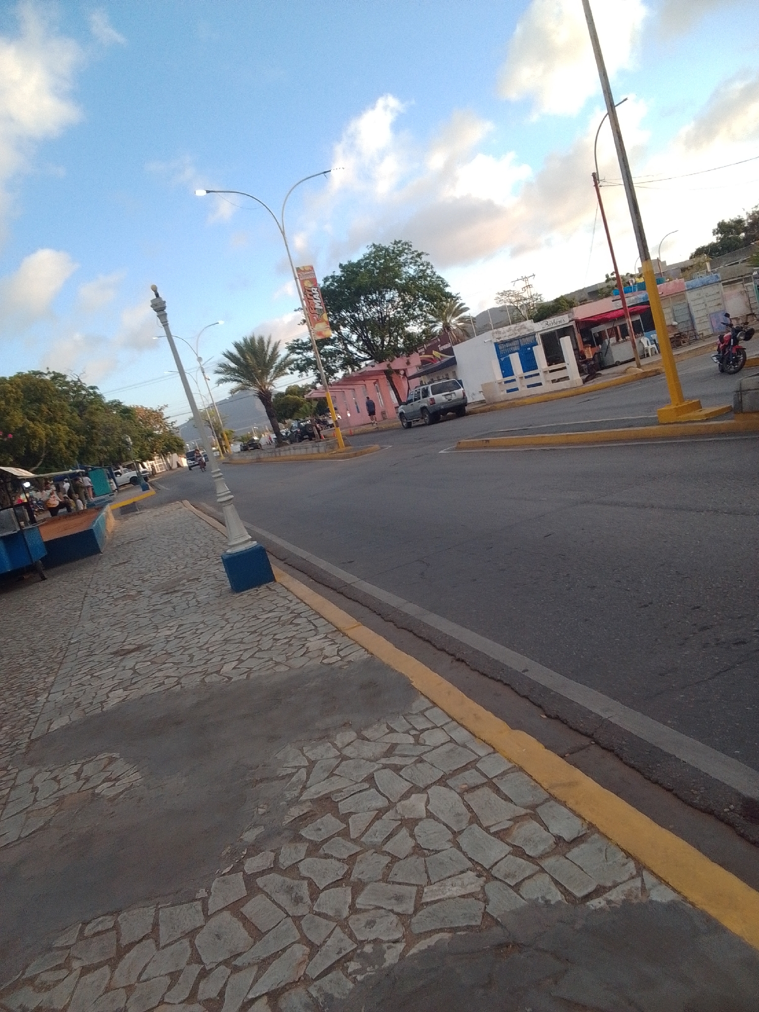 The image depicts a street scene in La Tagua, Venezuela. The perspective is from a sidewalk with a cobblestone surface, looking out towards a paved road. On the left side of the frame, several blue structures appear to be market stalls or vendor stands, with people gathered around them. A palm tree is visible in the background, along with trees lining the street. Across the road, there are buildings with colorful facades, some with awnings and signs. A yellow line marks the edge of the road. A silver SUV is parked on the side of the road, and further down, a motorcycle is visible, with a person on it. Streetlights are present along the sidewalk and road. The sky is blue with scattered white clouds.