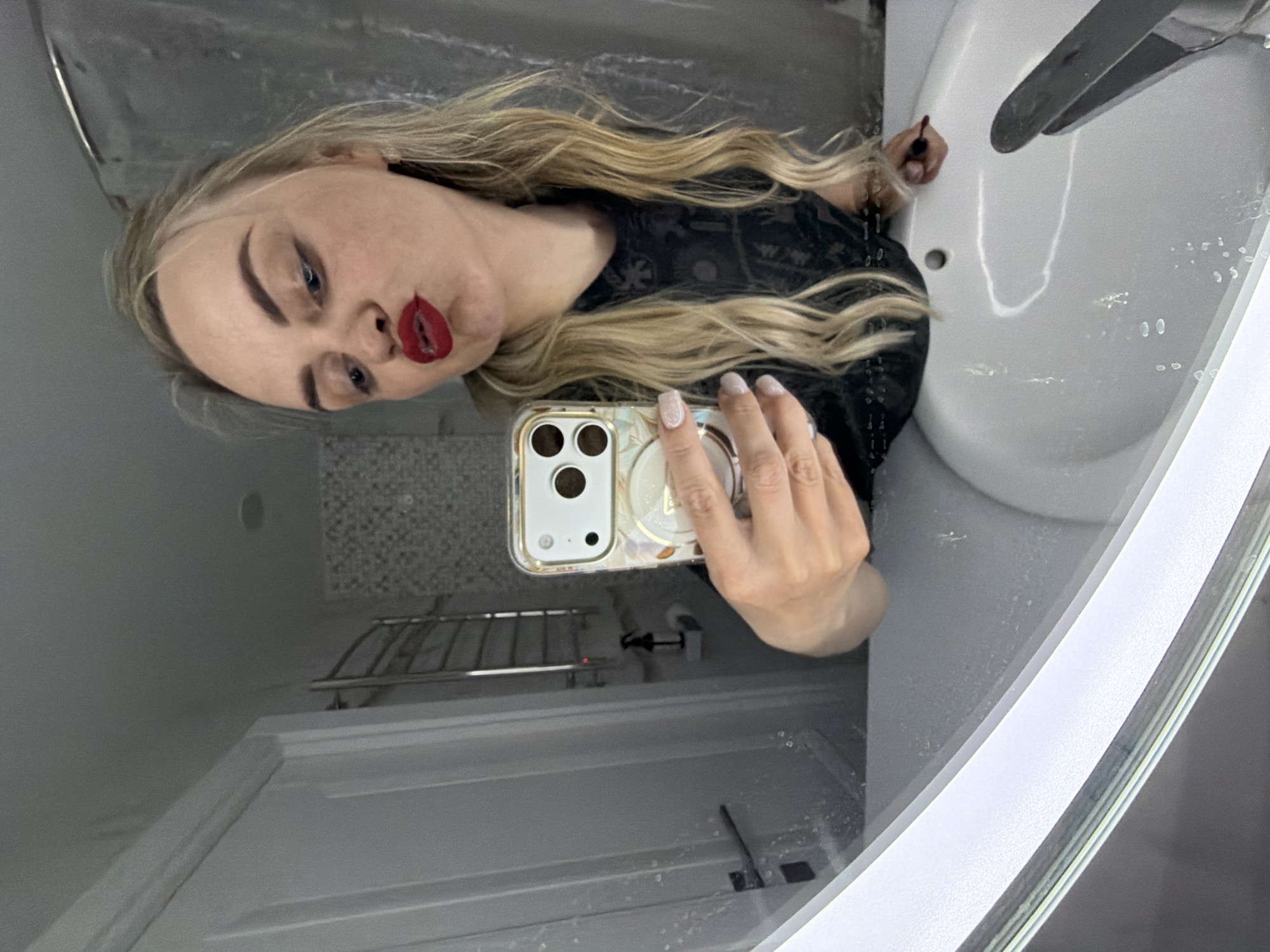 A blonde woman with red lipstick is taking a selfie in a bathroom. She is holding a smartphone with a protective case. The bathroom features a white sink with a chrome faucet, a mirror, a tiled wall, and a towel rack. The location is possibly a residential or commercial bathroom, but there is no specific information to determine if it is in Votkinsk, Russia.