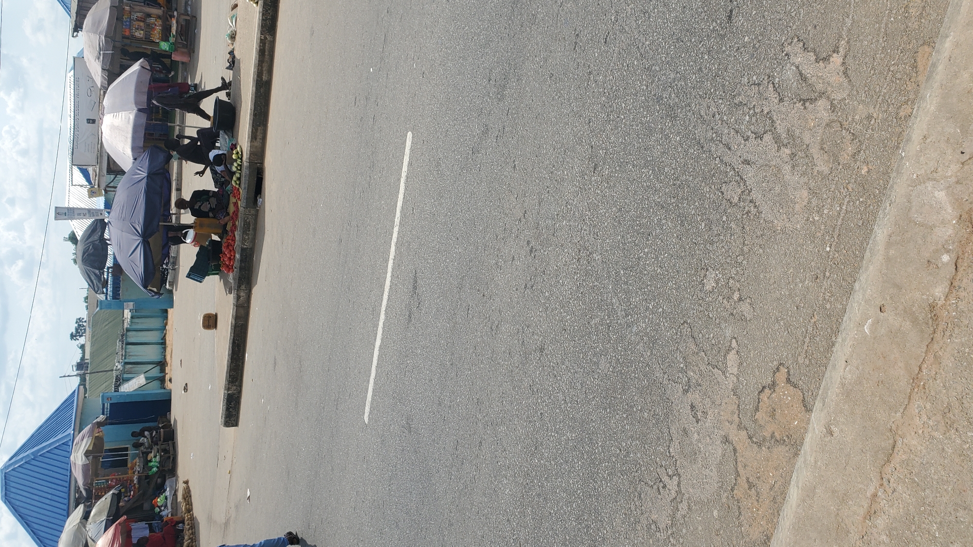 The image shows a street scene in Jalingo, Nigeria, with several vendors selling produce under umbrellas. In the foreground, a white line is painted on the asphalt road. In the middle ground, a group of people are gathered around stalls displaying fruits and vegetables, including tomatoes and cucumbers. Some individuals are seated while others are standing, and one person appears to be walking away from the scene. Umbrellas provide shade for the vendors and their goods. In the background, buildings and more market stalls can be seen. The sky is overcast.