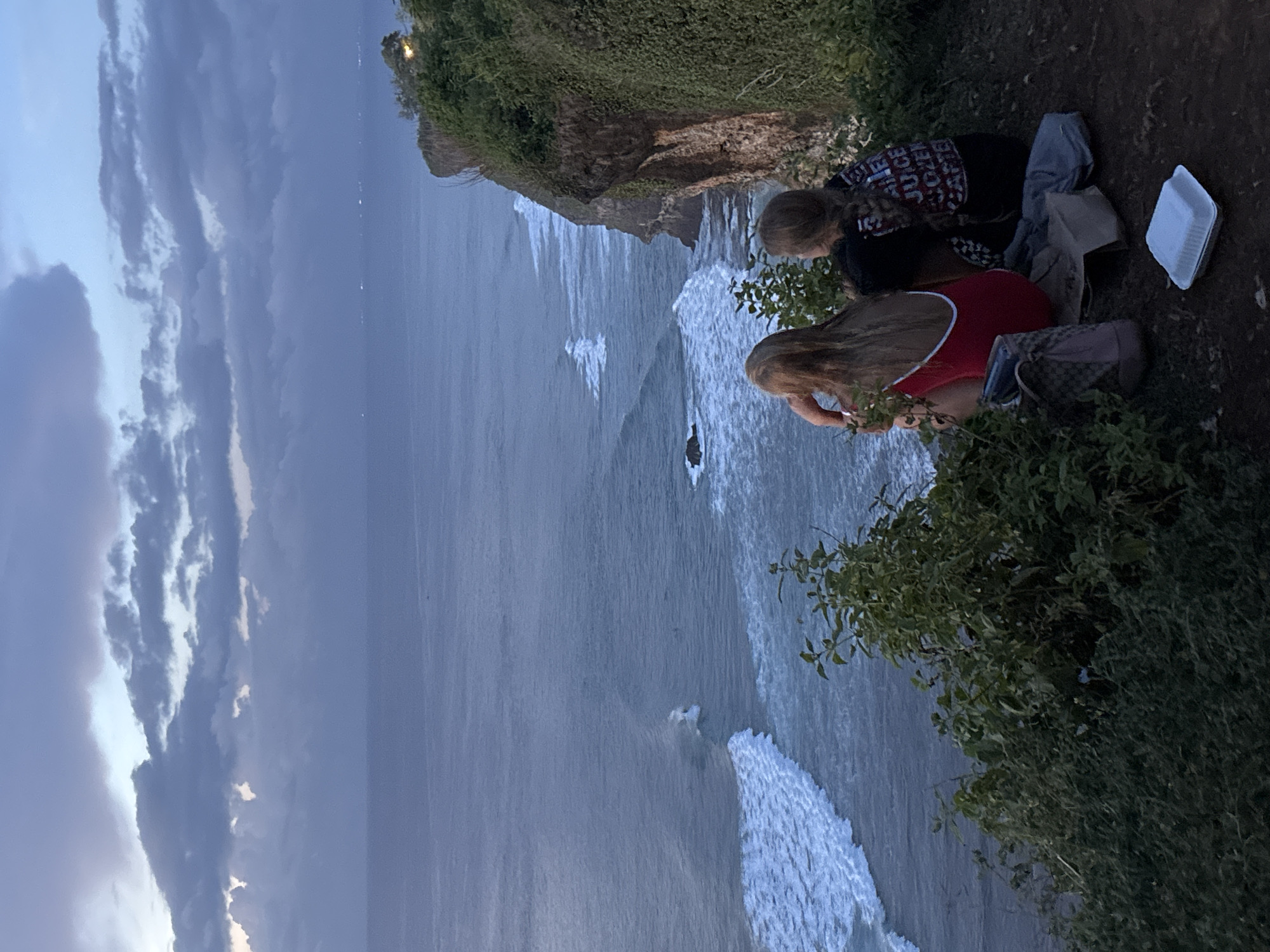 Two individuals are sitting on a cliff edge overlooking the ocean. The cliff is covered with vegetation. One person is wearing a black shirt with words printed on it, and the other is wearing a red top with white straps. A white styrofoam container, a gray bag, a purse, and a piece of clothing are near them. The ocean is visible below the cliff, with waves crashing against the rocks. The sky is cloudy. Based on the setting, this scene is likely in Uluwatu, Indonesia.