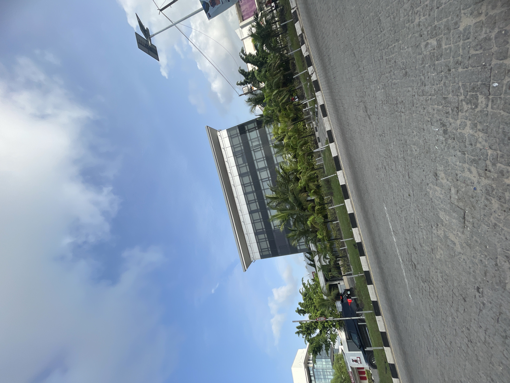 The image depicts a street scene in Ilado, Nigeria, angled from the ground upwards towards a cloudy sky. The road is paved with cobblestones and has a black-and-white painted curb. A modern office building is visible, surrounded by palm trees and other greenery. A traffic sign and a parked car are on the side of the road.