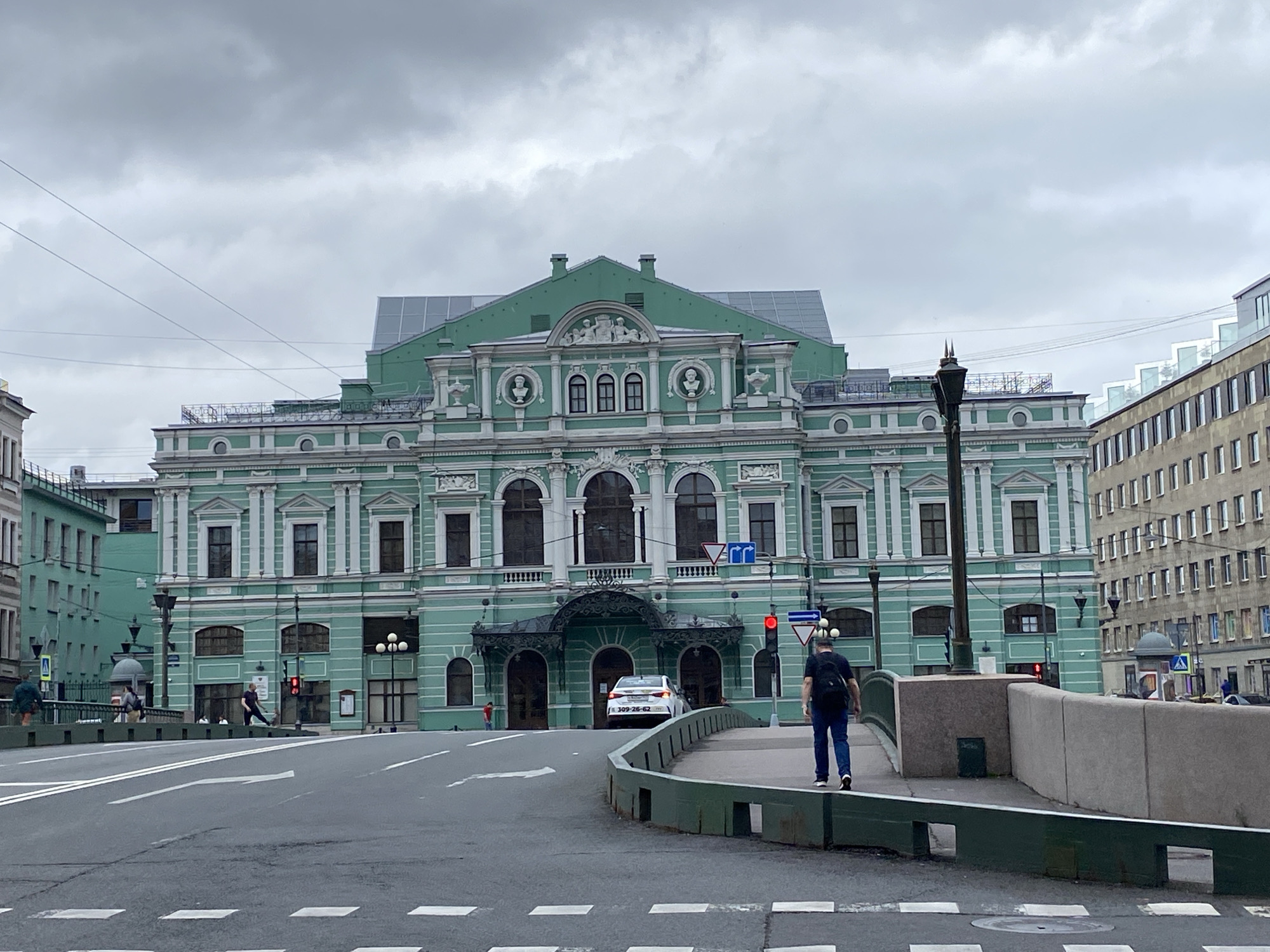 The image shows a wide street scene in Saint Petersburg, Russia, on a cloudy day. The dominant feature is a large, ornate, light green building with white decorative elements, likely a historic theater or opera house, with the Mariinsky Theatre being a strong possibility given its architectural style and color. In front of this building, a white car is visible. A man with a black backpack walks away from the camera on a pedestrian walkway that curves to the right, with a green metal railing bordering it. Across the street, other buildings and traffic lights are present. The road in the foreground has clear white lane markings and pedestrian crossings. Power lines are visible crisscrossing the sky.