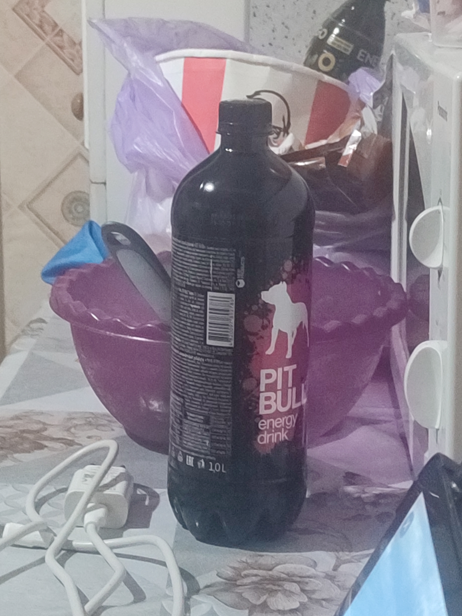 The image shows a domestic indoor scene, likely a kitchen or utility area, with various objects cluttering a tabletop. A large, dark bottle of "PIT BULL energy drink" is prominently displayed in the foreground. To its left, a purple bowl contains a blue funnel and a kitchen utensil. In the background, a white microwave is visible, along with a crumpled purple plastic bag and a portion of a cardboard container. A white electrical charger and cable are also present on the table. The overall impression is one of disarray and everyday life. The location in Chernivtsi, Ukraine is not discernible from the visual cues in the image.