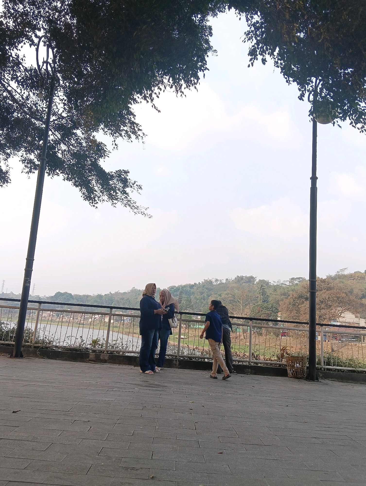 In Padalarang, Indonesia, on a paved walkway overlooking a body of water, four individuals are present. Two women, wearing hijabs and dark blue tops, stand together. One is holding a phone. To their right, a young boy in a blue shirt and light-colored pants walks away from the camera, while another individual, whose gender is not clearly discernible, stands next to him, facing the water. A metal railing separates the walkway from the water's edge, where vegetation is visible. Two tall lampposts with decorative tops are positioned on either side of the walkway, and trees with dark leaves arch over the scene. In the background, a hillside with scattered buildings and foliage rises against a hazy sky.