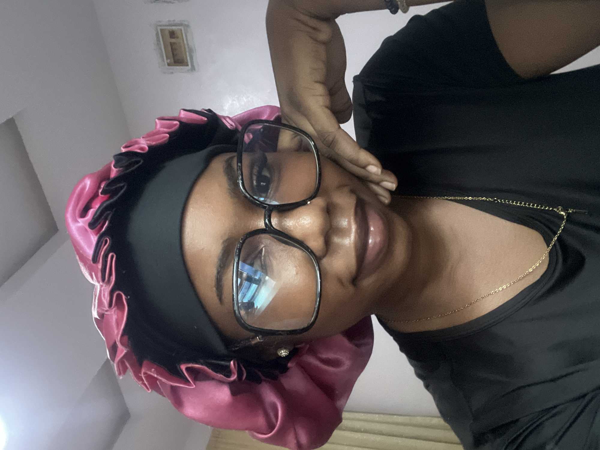 A close-up, chest-up portrait taken indoors in Rafin Par, Nigeria, depicts a young adult female with dark skin. She is looking directly at the camera with a slight, closed-lip smile. She wears large, square-framed black glasses, the lenses of which reflect a bright overhead light source. Her hair is covered by a black fabric headband, over which a voluminous, ruffled, glossy pink satin bonnet with a black trim is worn. A small, round, silver-colored stud earring is visible in her left earlobe. She is dressed in a plain black short-sleeved top and a thin gold-colored chain necklace. Her left hand is raised to her face, with fingers lightly curled and resting against her right cheek and jawline. The background consists of light-colored, plain interior walls. A light-colored curtain or blind is partially visible on the far left, and a recessed, rectangular object (possibly a framed vent or decorative item) is present on the right wall. The ceiling is also light-colored, and the scene is well-lit.