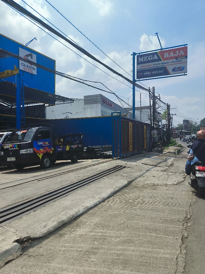 The scene shows a street in Ngamprah, Indonesia (latitude: -6.868, longitude: 107.524). There is a blue truck parked on the right side of the street. The truck has a sign on the side that says "GASCOOL". There are several power lines overhead. There is a blue and yellow gate in the background. To the left of the truck, there is a sign that says "Cilegon Steel". On the right side of the street, there is a sign that says "MEGA BAJA". In the distance, there are more buildings, trees, and a person riding a motorcycle. The street is paved with concrete, and there are some cracks in the concrete. The sky is blue and there are some clouds. The image shows a typical scene of a street in a developing city in Indonesia. There are signs in Indonesian and English. There are several different kinds of vehicles on the street. There are many power lines in the image, which may be a sign of a city with a high population density.
